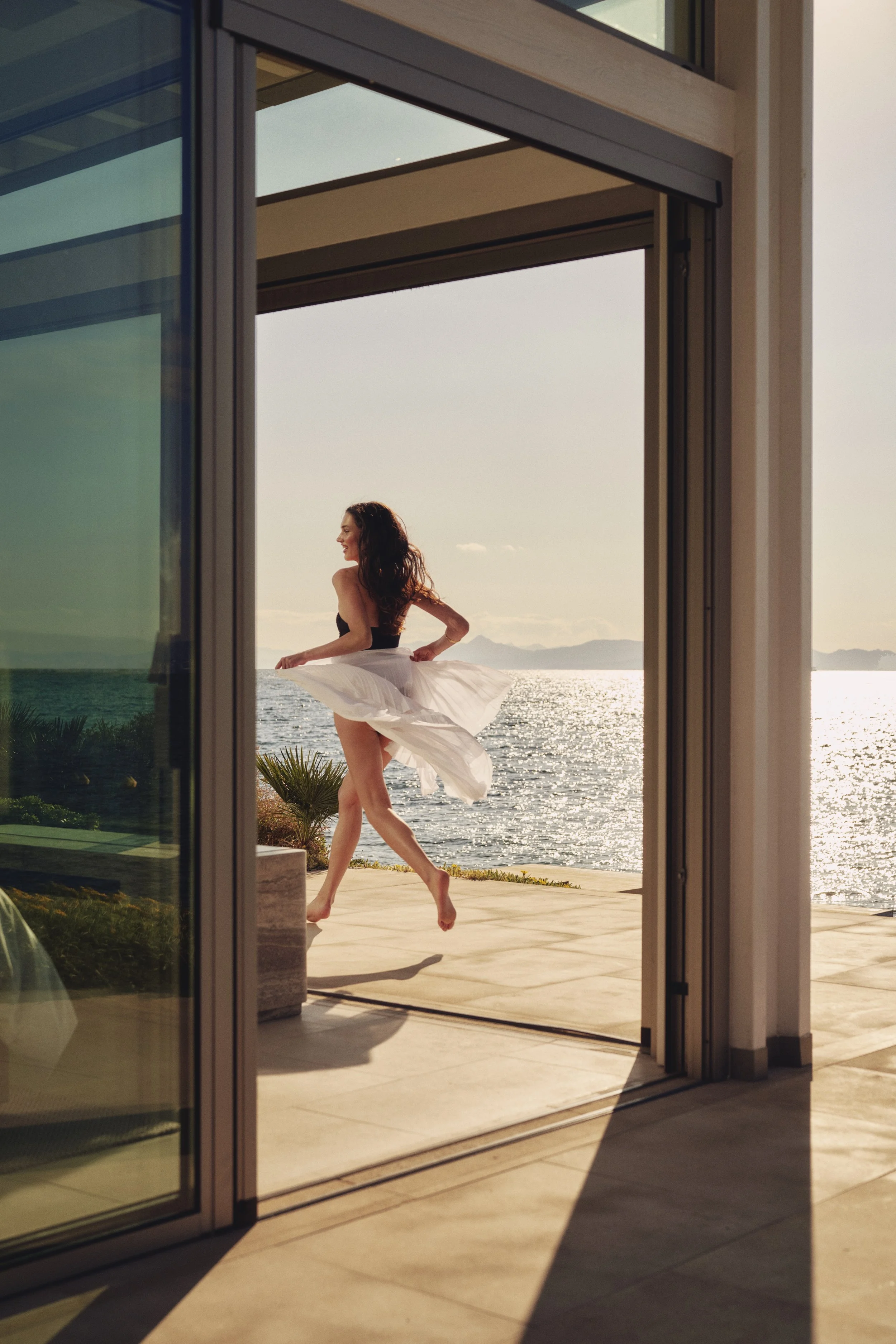 Woman dancing on a balcony overlooking the ocean. Photo taken from inside a building through open glass doors.