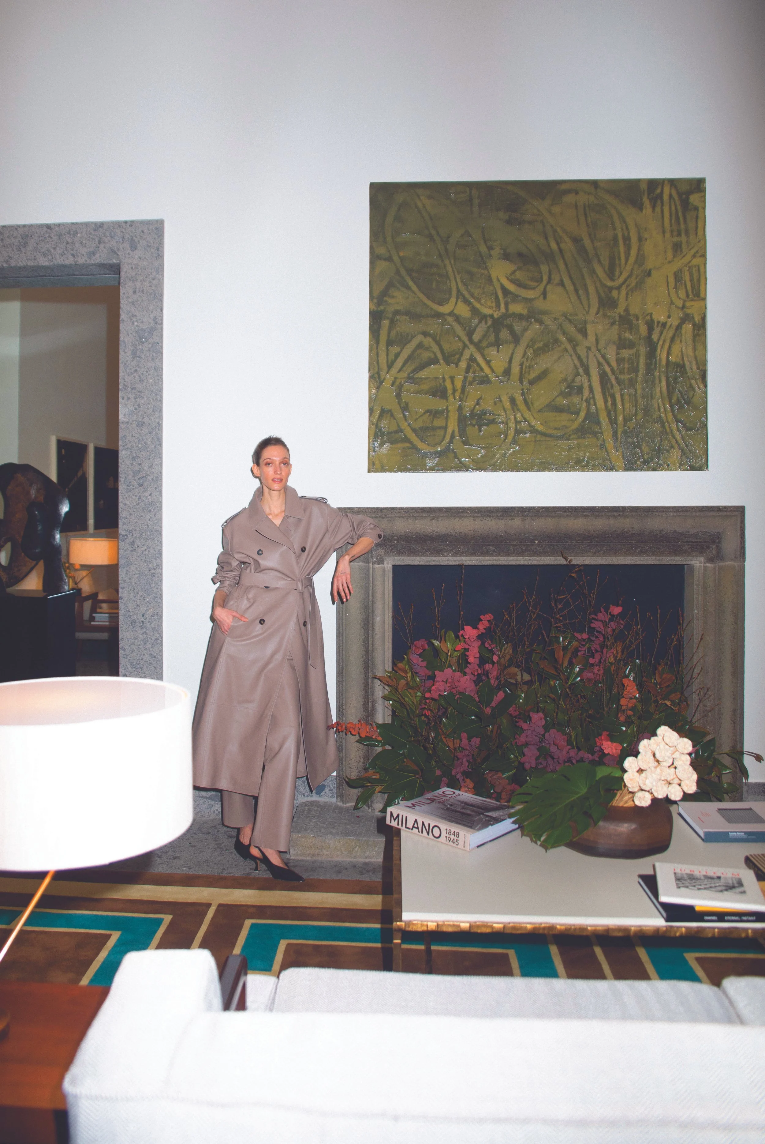 A woman in a beige trench coat and black high heels standing next to a fireplace with a large abstract yellow and black artwork above it. There are flowers and books on a table in front of the fireplace, with a colorful geometric rug on the floor. Pa