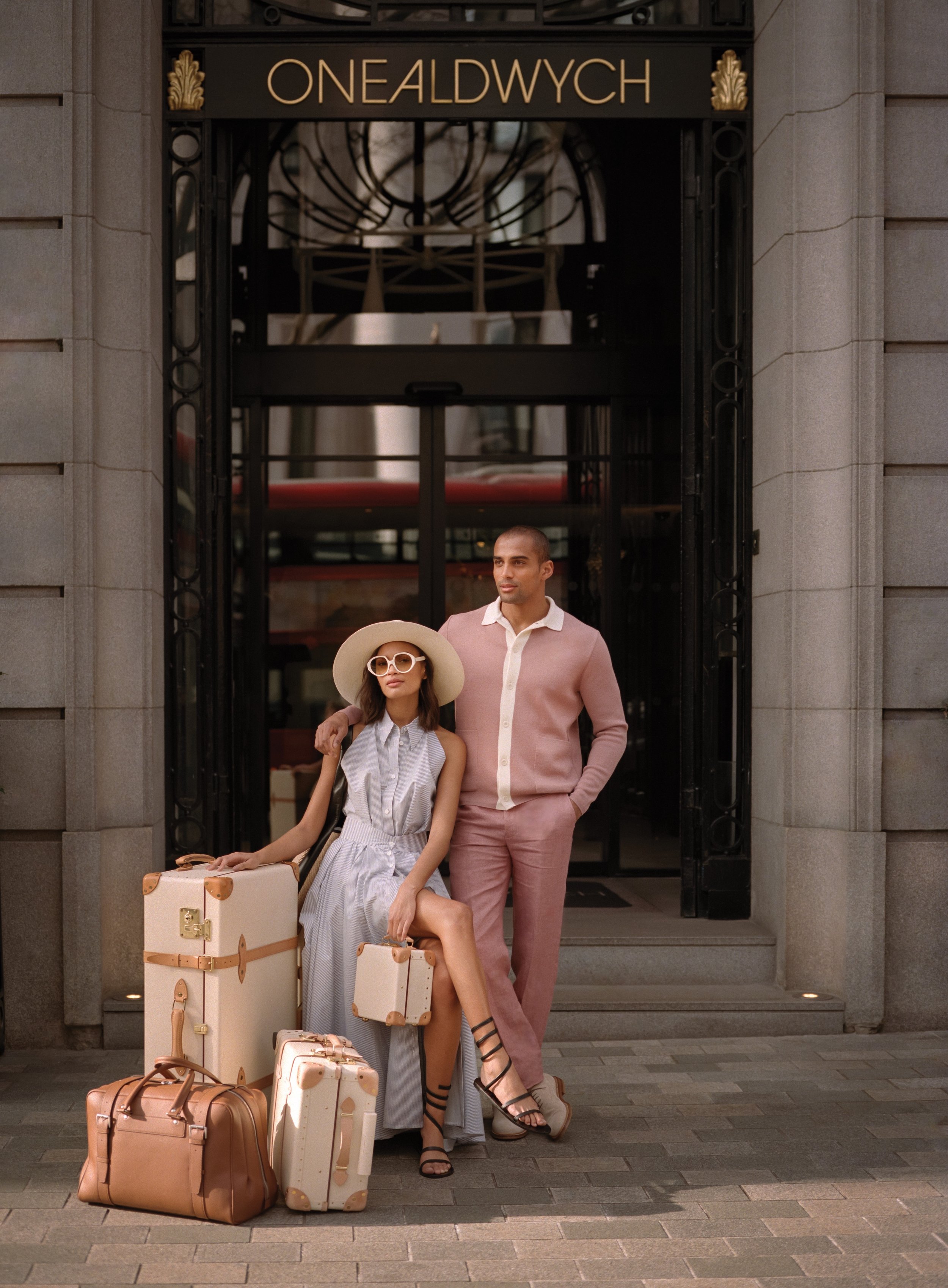 A stylish couple stands with luggage in front of a building entrance, which has a black awning with gold lettering that reads 'ONEALDWYCH.' The woman is sitting on a suitcase, wearing a light blue dress, large sunglasses, and a wide-brimmed hat. The 