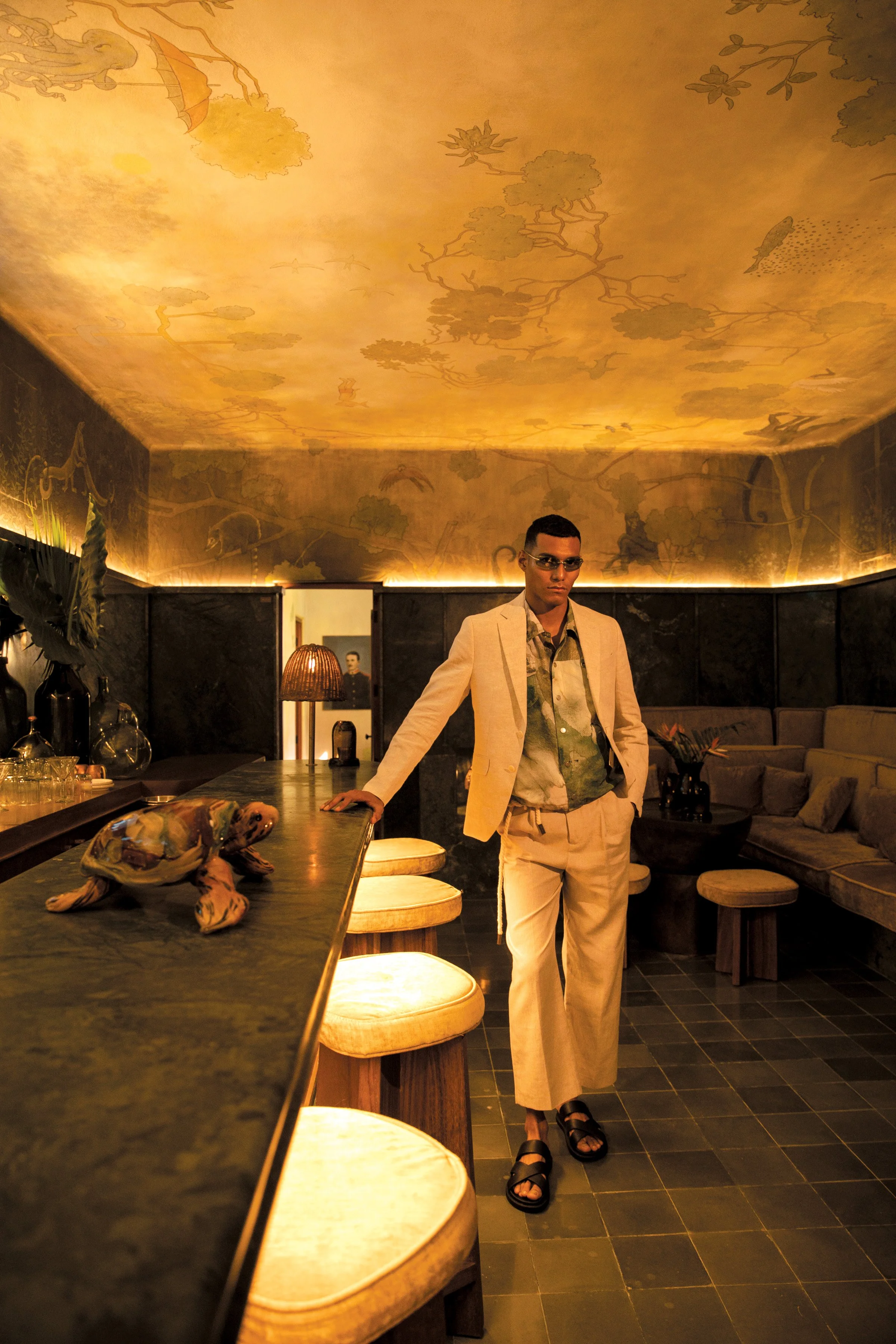 A man wearing a beige suit, camouflage shirt, sunglasses, and sandals standing in a dimly lit lounge or bar with a mural of trees and animals on the ceiling and a black marble wall in the background. A wooden turtle sculpture is on the bar counter.