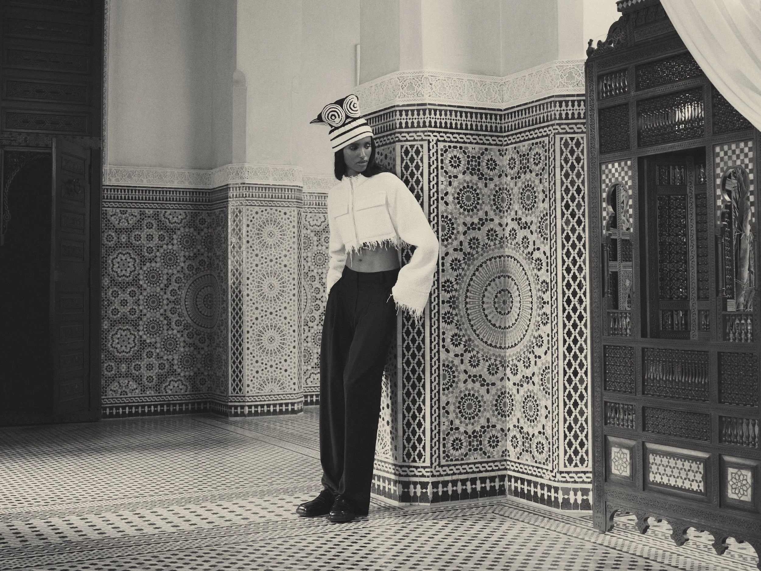 A woman in a striped hat and cropped hoodie standing in an ornately decorated room with Moroccan-style mosaic tile walls and intricate wooden doors.