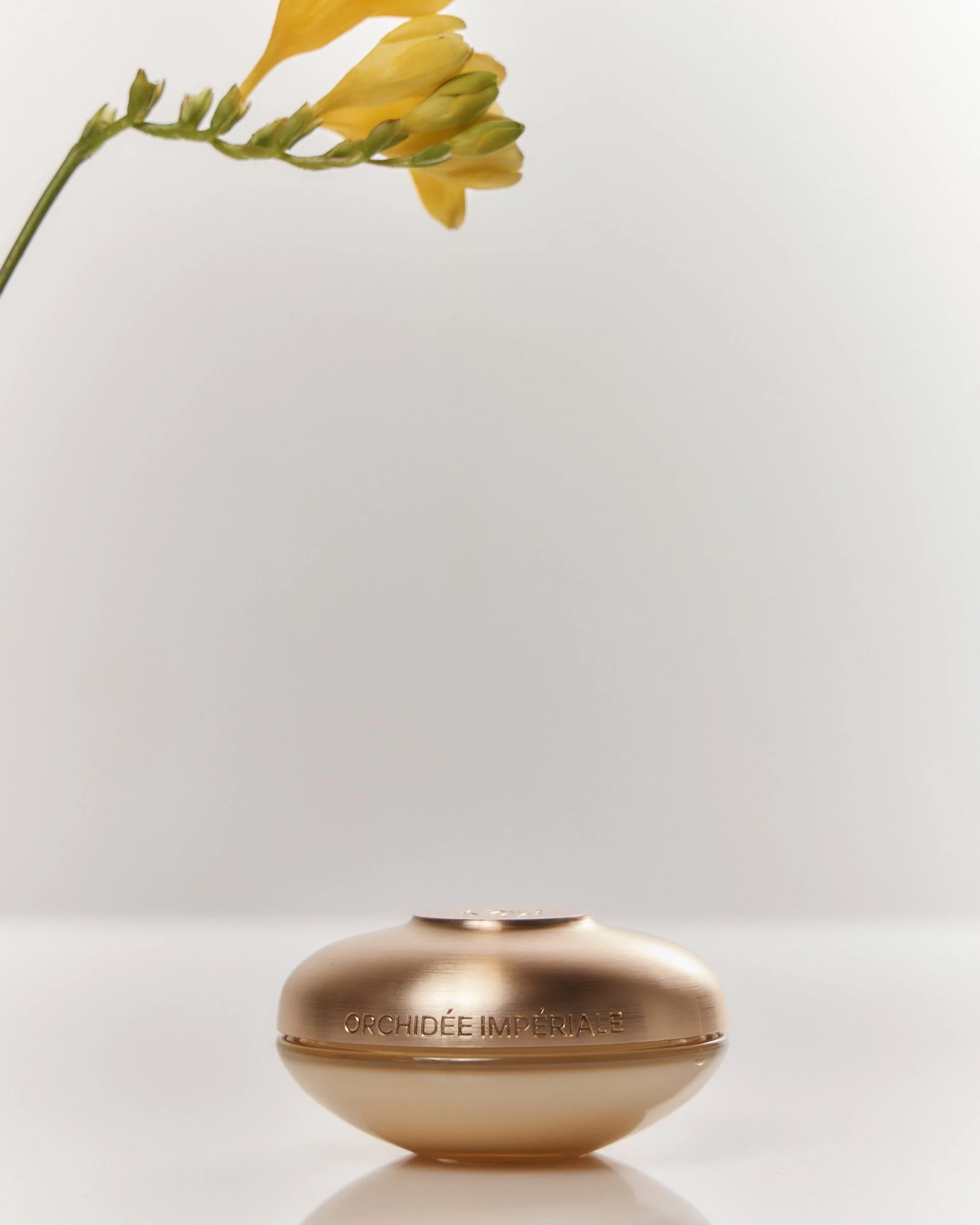 A small, elegant gold vase with the words 'Orchidee Impériale' engraved on it. The vase contains yellow orchid flowers with green buds. The background is plain white, emphasizing the minimalistic and luxurious appearance of the arrangement.