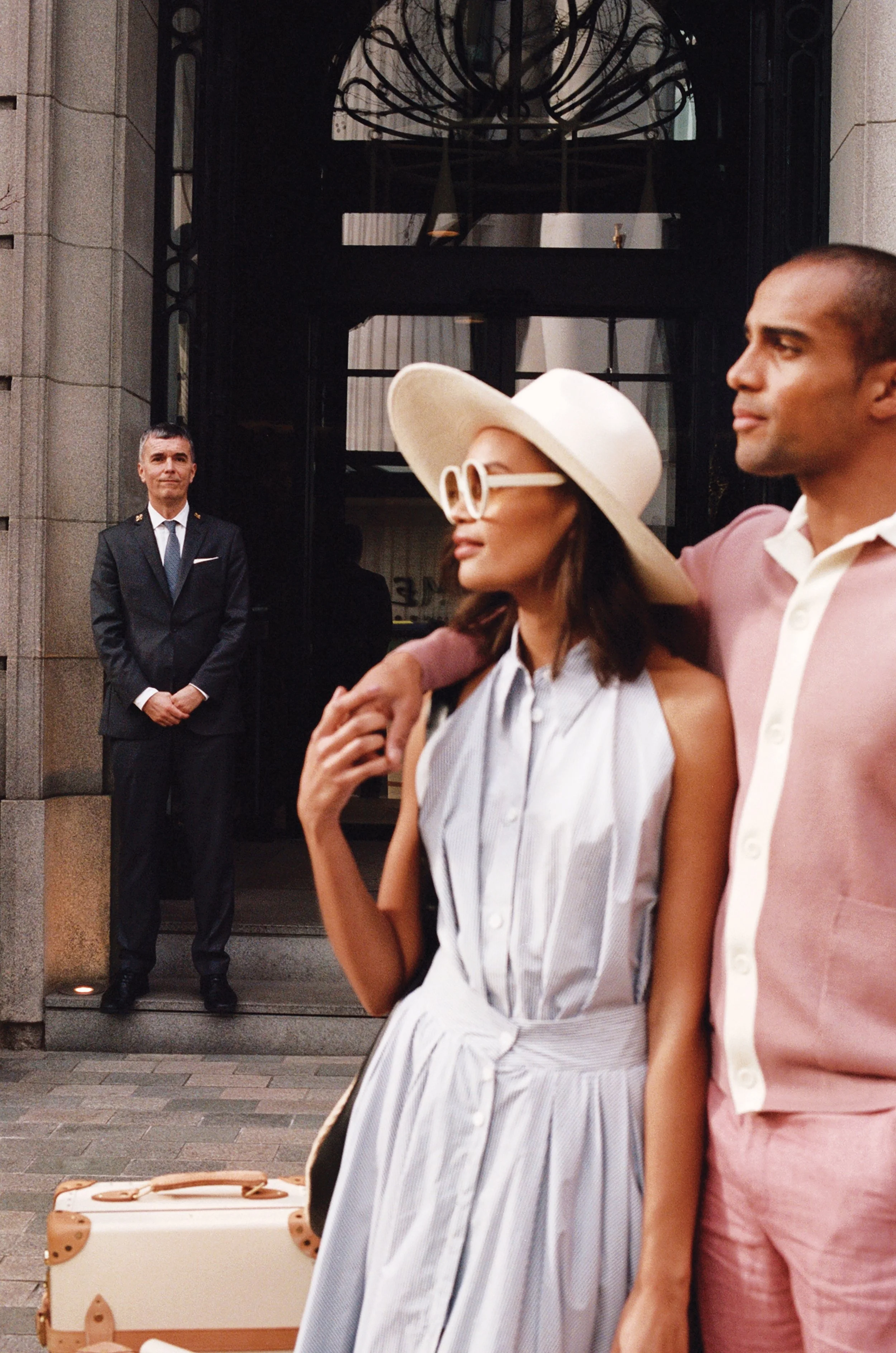 A man in a dark suit and tie standing with hands clasped outside of a building, while a woman in a light striped dress, large white hat, and sunglasses, and a man in a pink shirt walk past in the foreground.