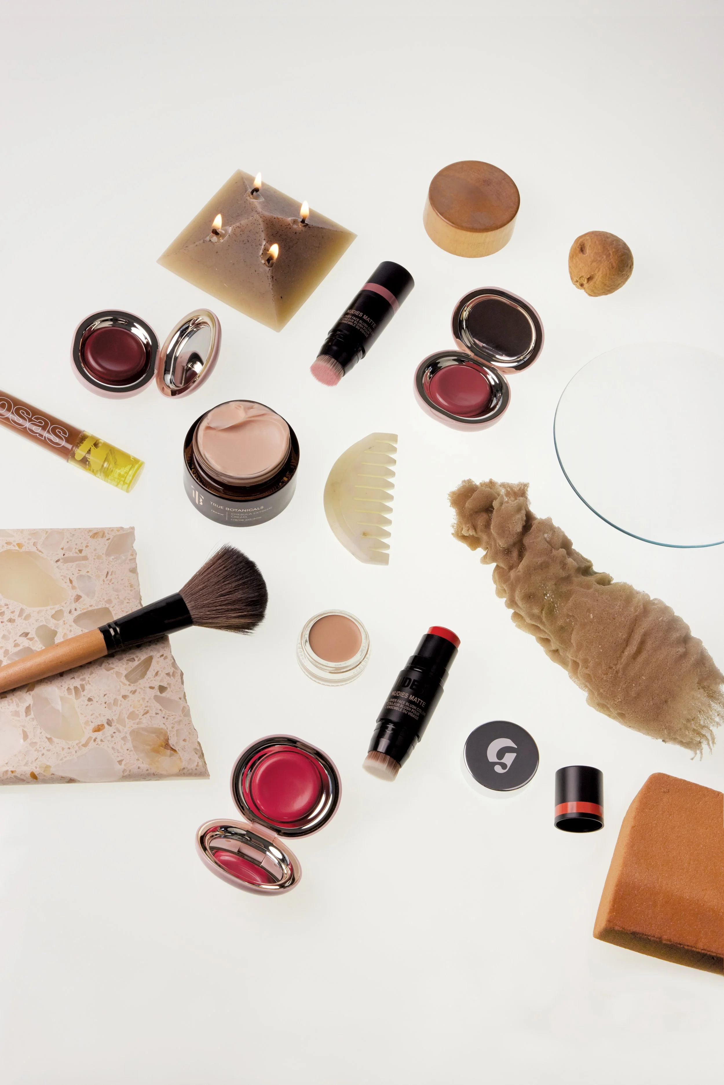 Beauty products on a white surface, including makeup, skincare, brushes, and natural elements like a candle and rocks.