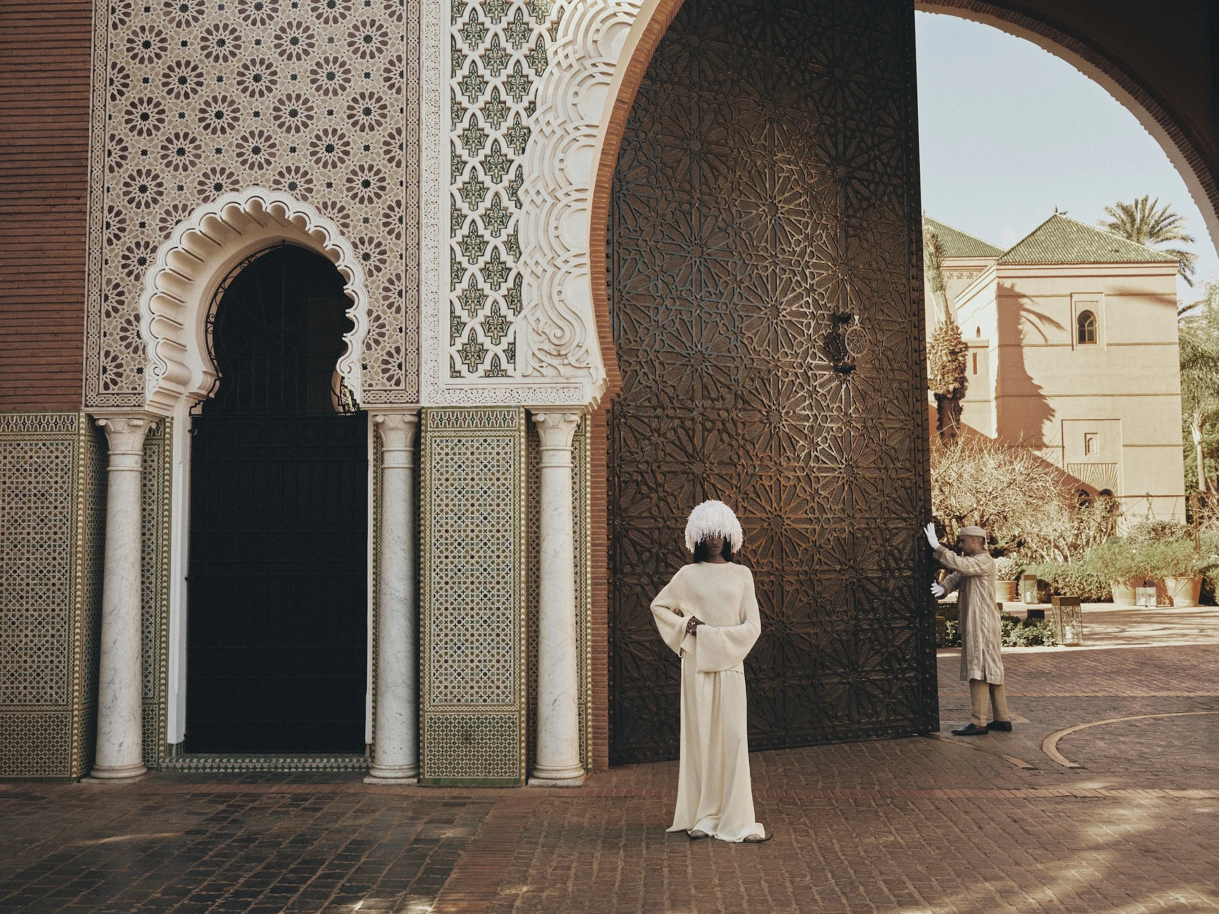 A man and a woman dressed in traditional Moroccan clothing standing on the sidewalk outside an ornate building with intricate tile and woodwork designs. Palms and other buildings are visible in the background.