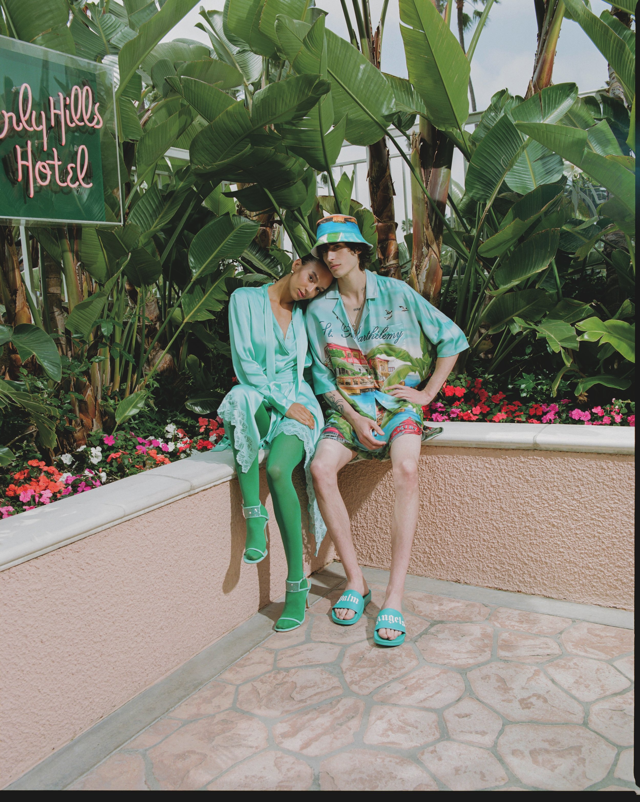 Two young models sitting outdoors in front of lush green banana plant leaves, one with a head covering, both dressed in pastel-colored outfits, with a pink flower bed and a neon sign that reads 'North Hills Hotel' in the background.