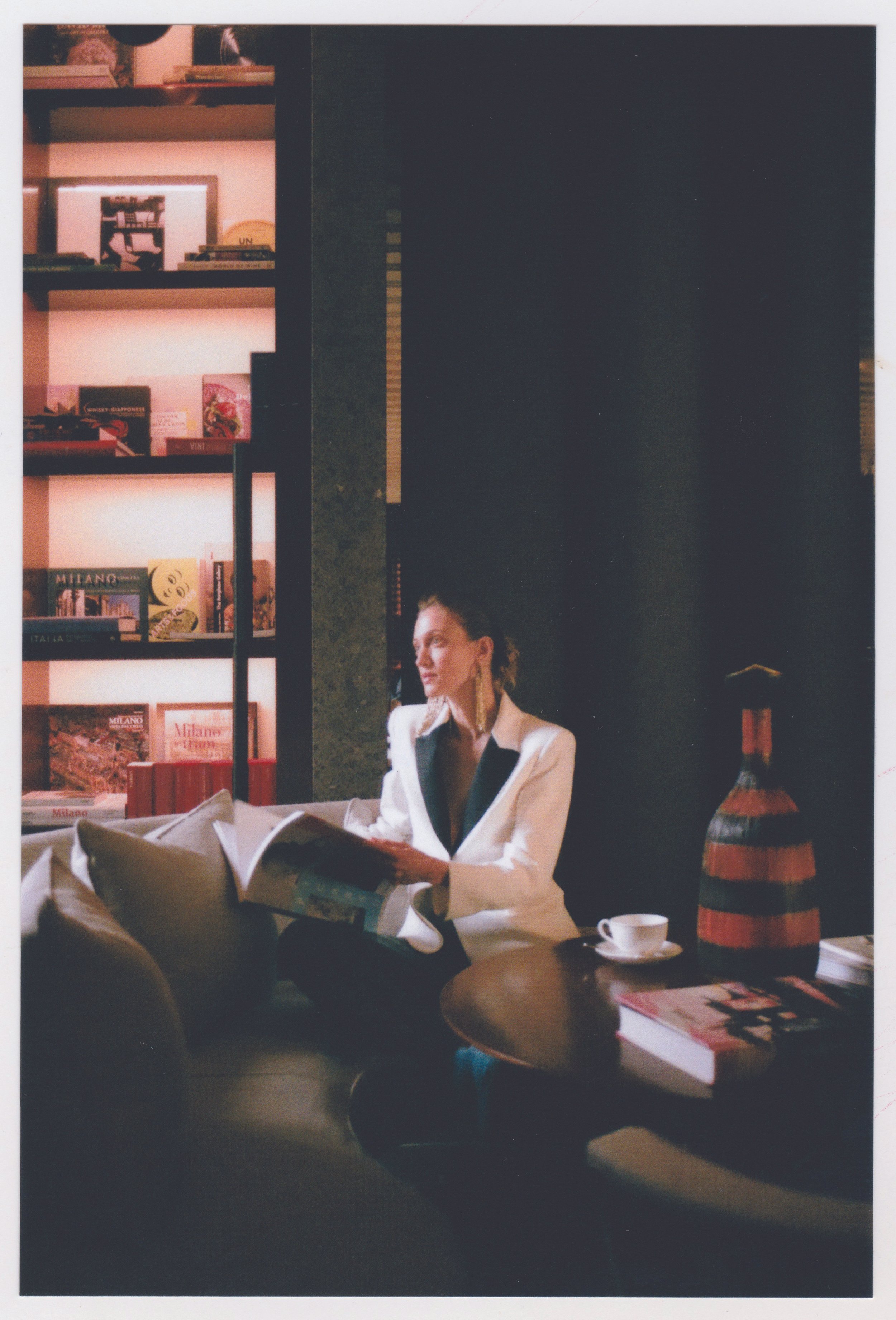 A woman sitting at a table with a cup of coffee, surrounded by books and magazines, in a cozy room with a dark wall and a bookshelf in the background.