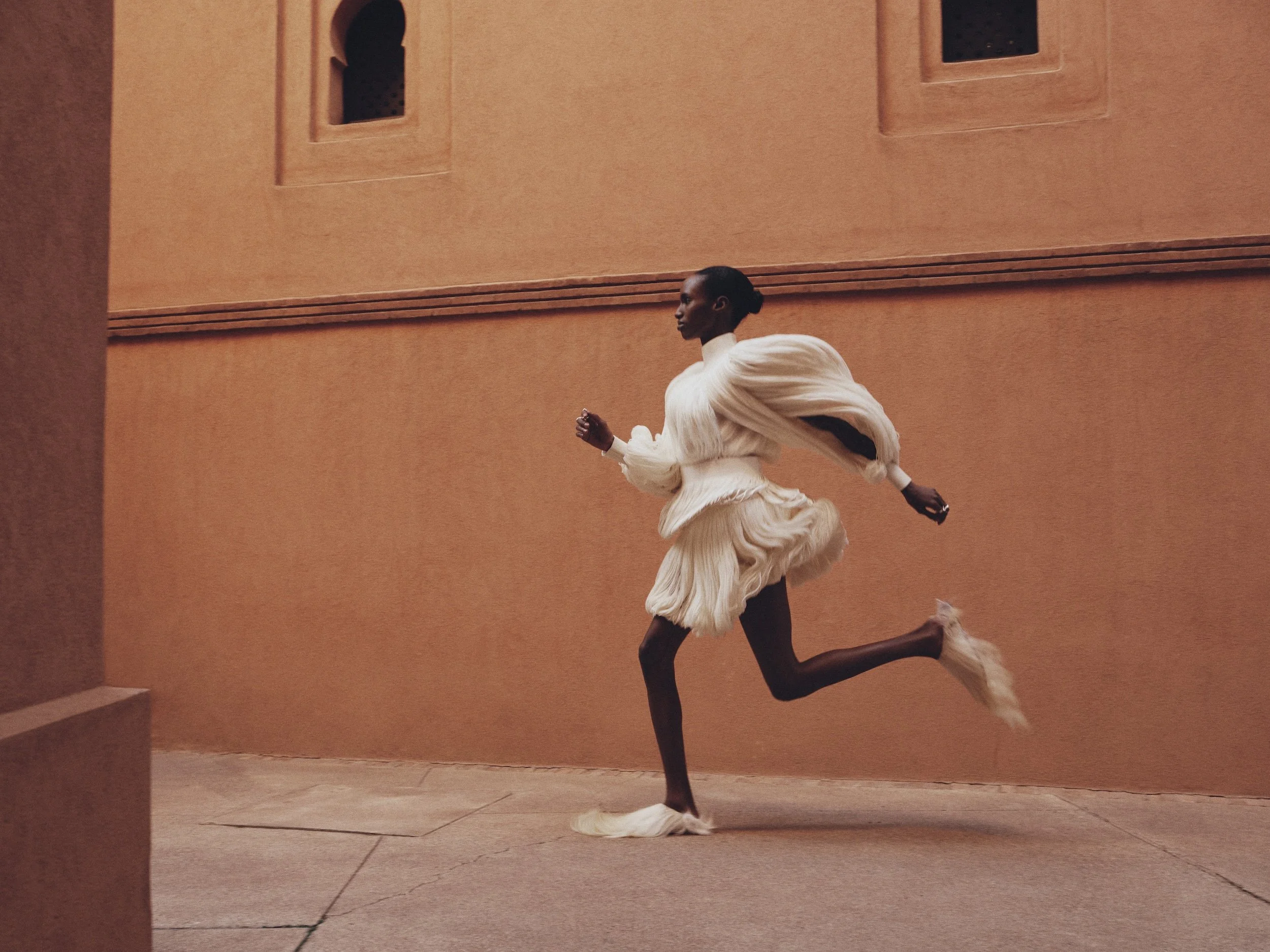 A performer runs on a stage with a peach-colored wall background, wearing a cream-colored outfit with flowing fabric. The stage floor is tiled.