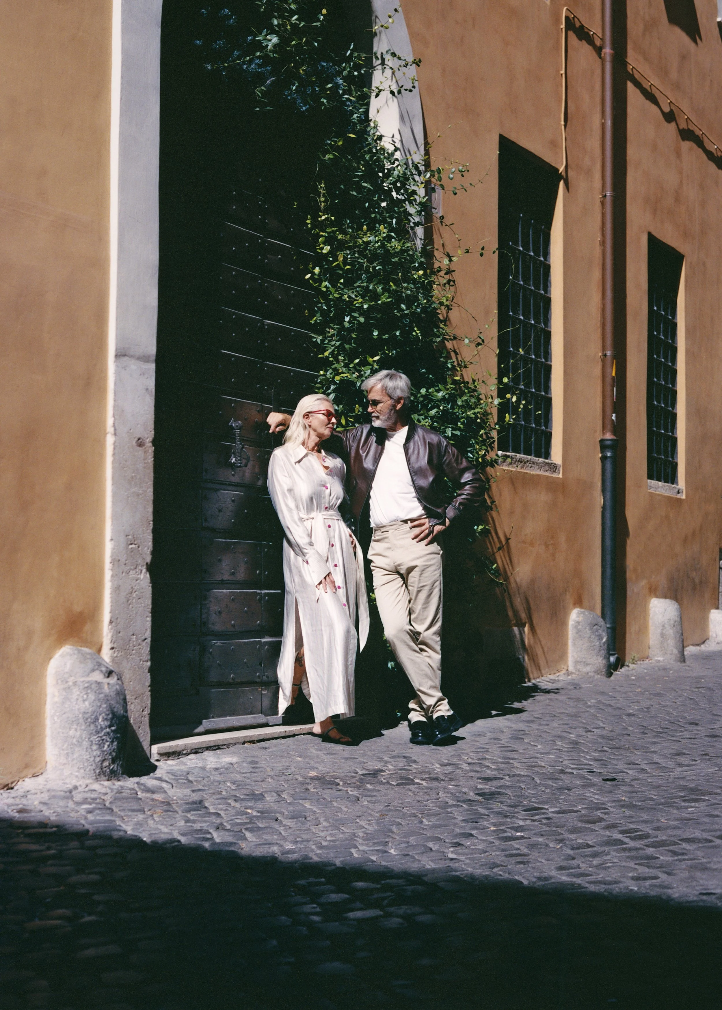 A man and woman are standing close together on a cobblestone street in front of a large wooden door and a building with an orange wall. The woman has long blonde hair, is wearing glasses, a long white dress, and is looking at the man. The man has gra