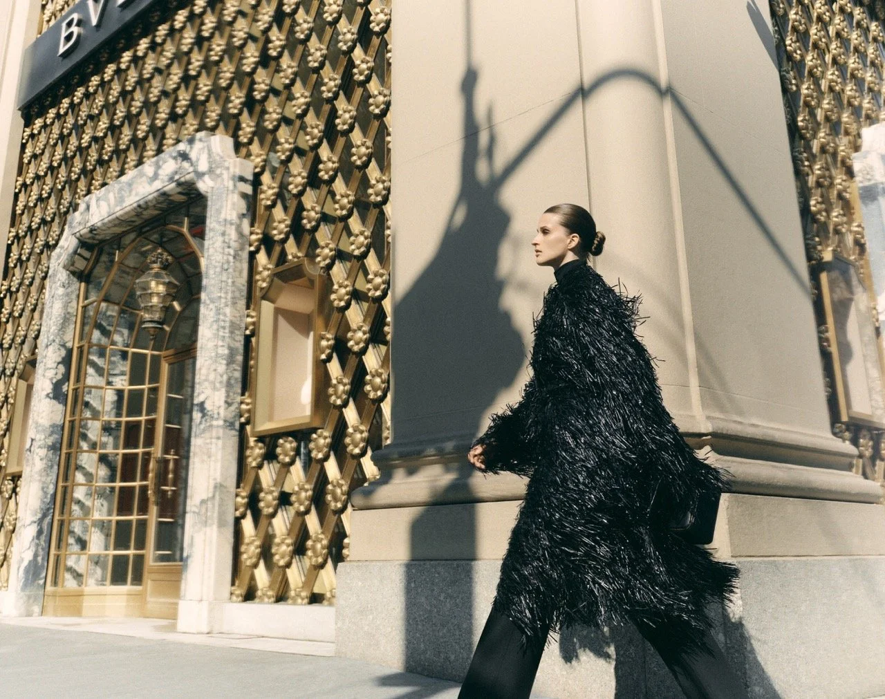 A woman dressed in a black, textured coat walks past a building with gold decorative elements and marble accents, casting a shadow on the wall.