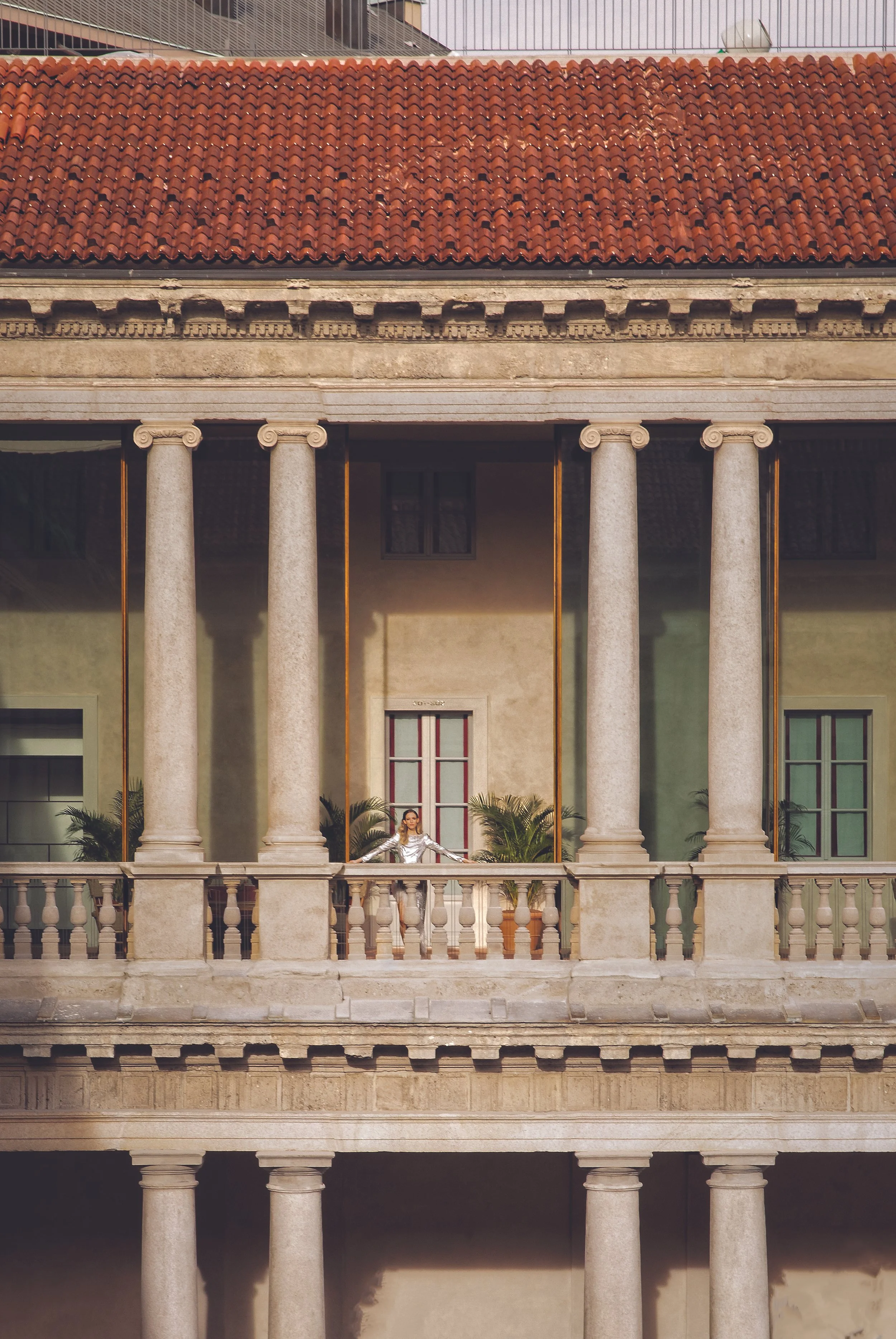 A woman in a shiny silver outfit stands on a balcony with four large stone columns, in front of a building with a red tile roof and classical architectural details.