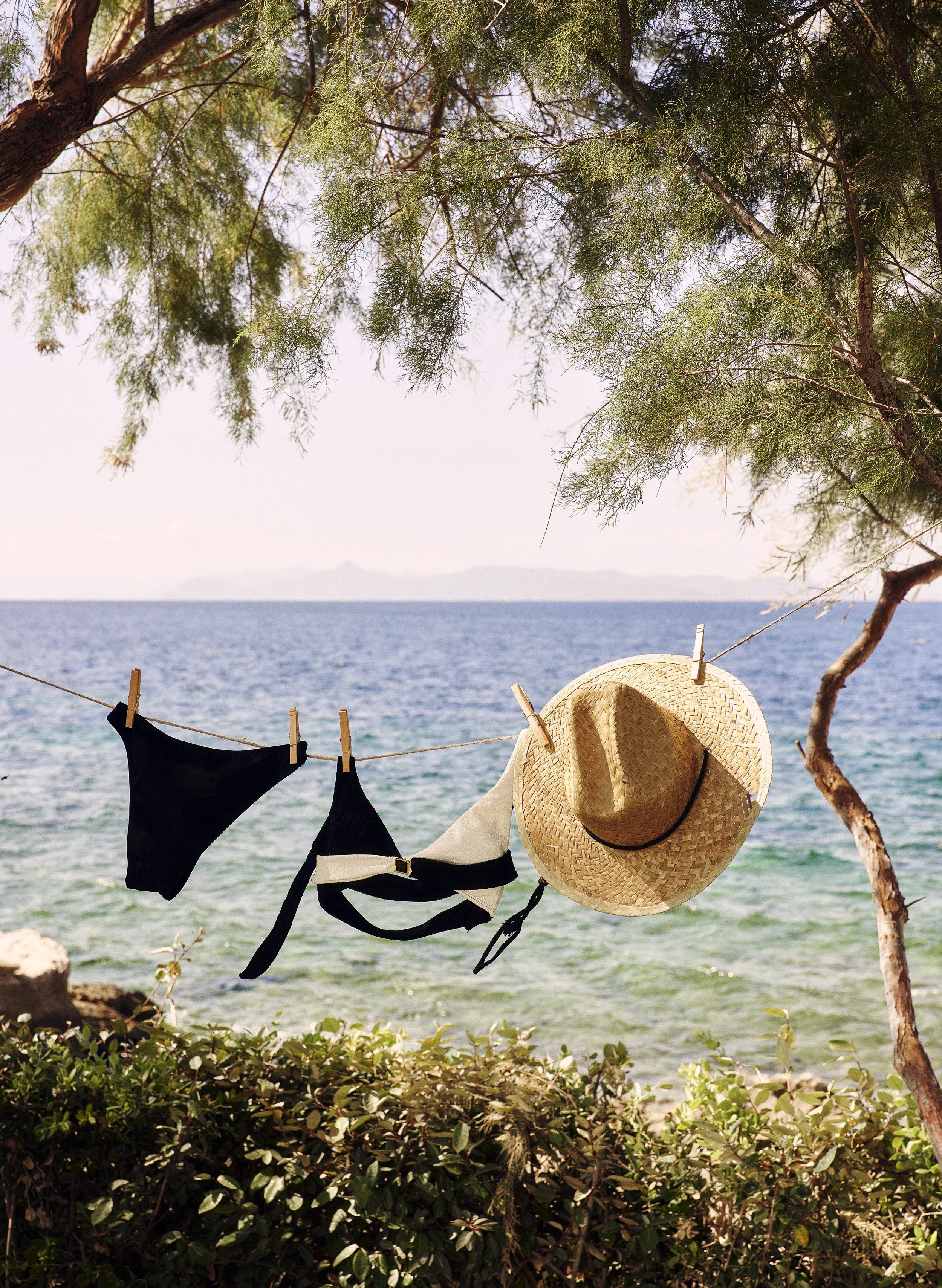 Clothes and accessories hanging on a clothesline near the beach, including a black bikini top and bottom, and a straw hat with a black band.