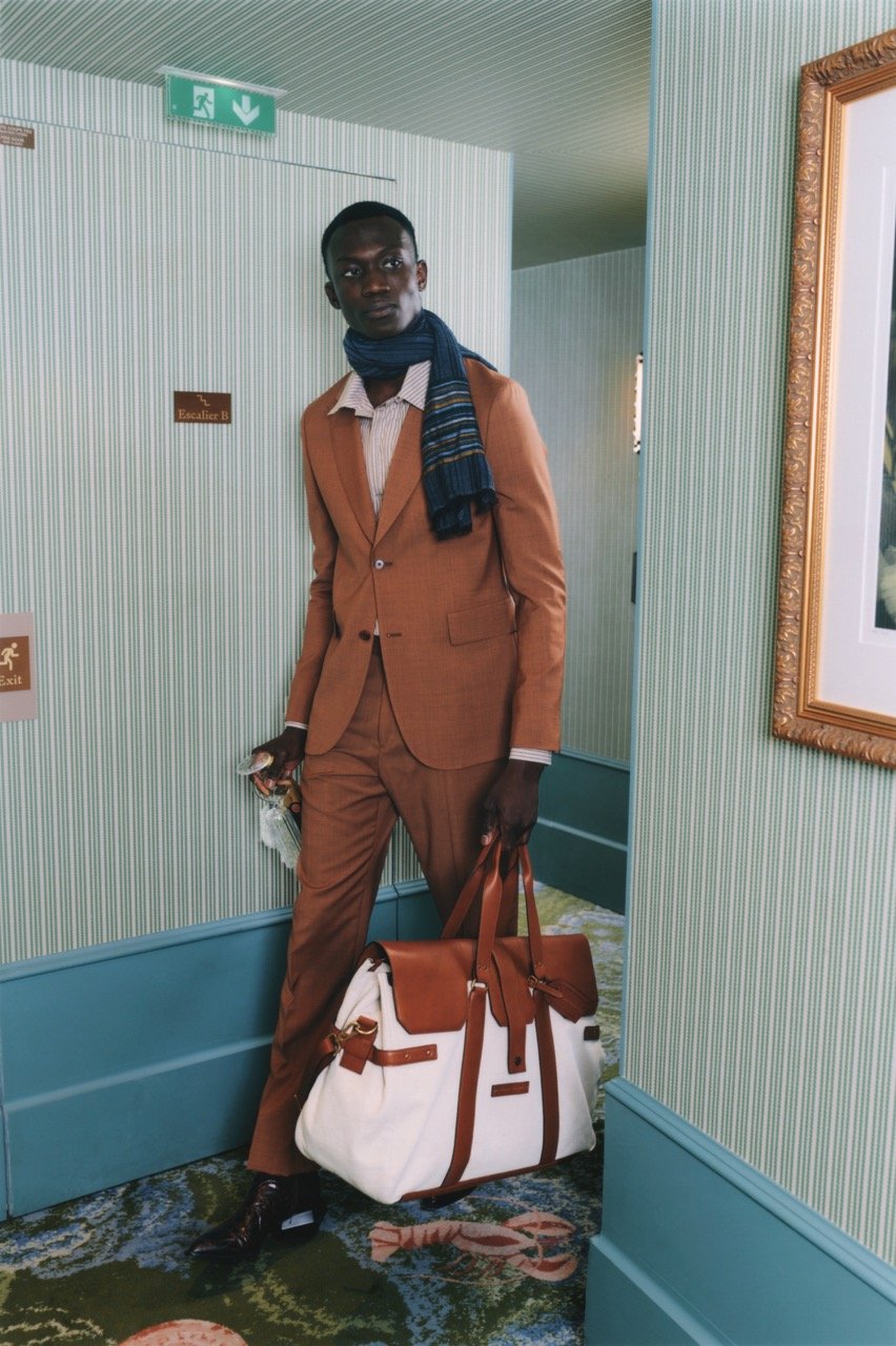 A young man wearing a brown suit and blue scarf, standing in a hallway, holding a large white and brown duffle bag, with patterned carpet and framed artwork on the wall.