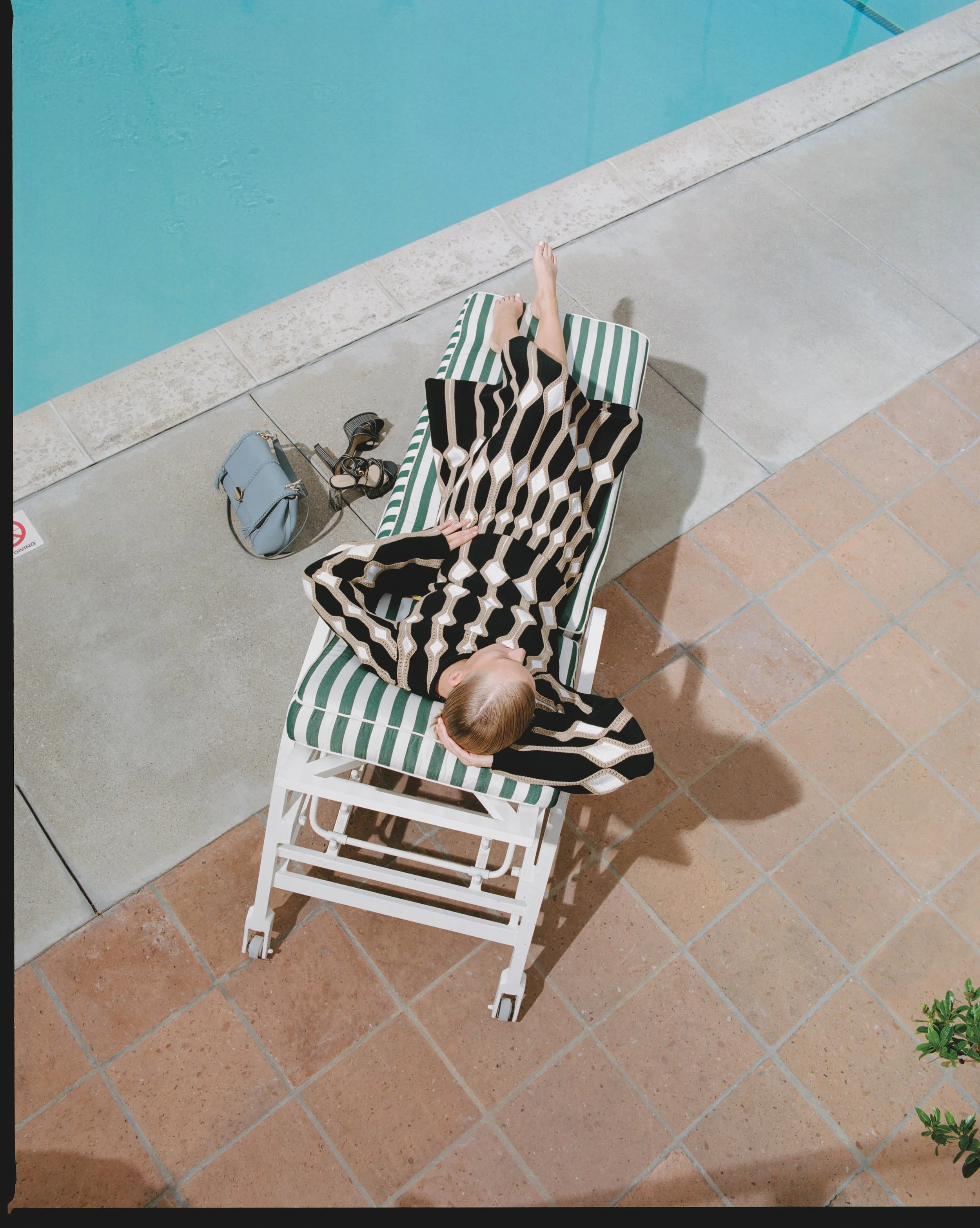 A person is lying on a green and white striped pool lounger next to a swimming pool, with items including a light blue purse, black sunglasses, and black sandals nearby. The person is wearing a black, white, and beige geometric patterned outfit, with