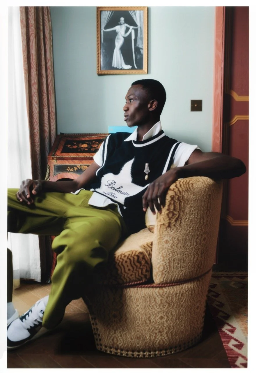 A person with dark skin and short hair sitting in an elegant living room, wearing a black and white Balmain Paris vest, olive green pants, and white sneakers, looking to the left. Behind them is a framed black and white photo of a woman in a formal d