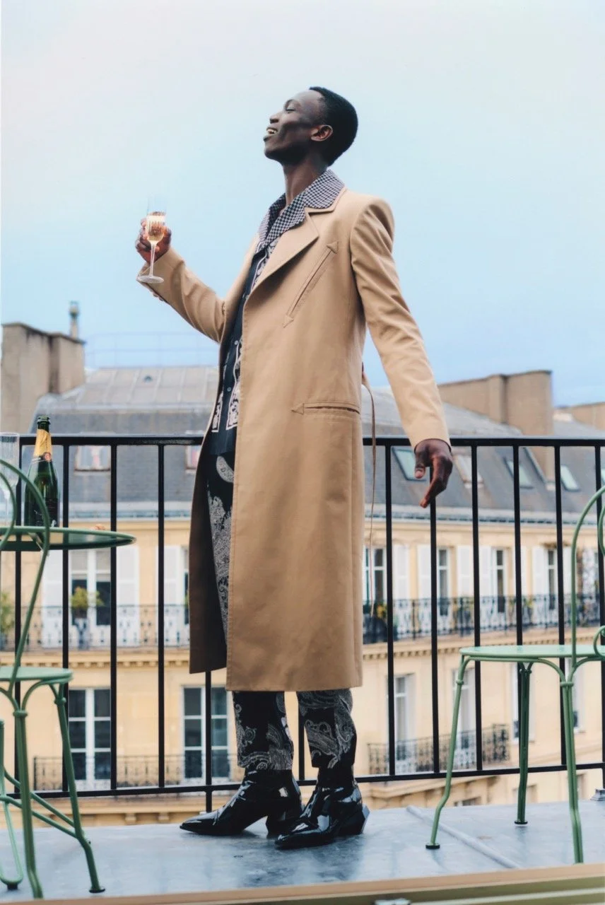 A person standing on a balcony holding a glass of champagne, wearing a stylish beige trench coat, patterned pants, and shiny black shoes, with a cityscape in the background.