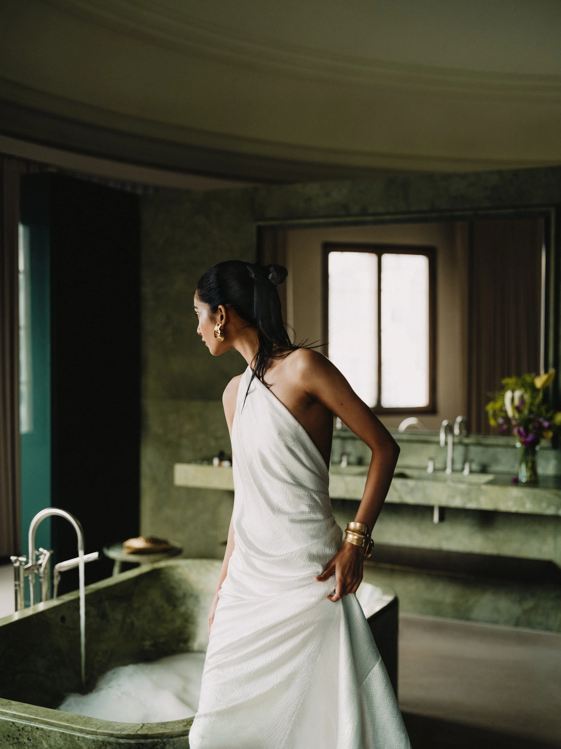 A woman with black hair styled in a bun, wearing large gold hoop earrings and a white towel wrap, standing in a bathroom with green marble countertops and a sink, near a window with curtains, and a vase with flowers on the counter.
