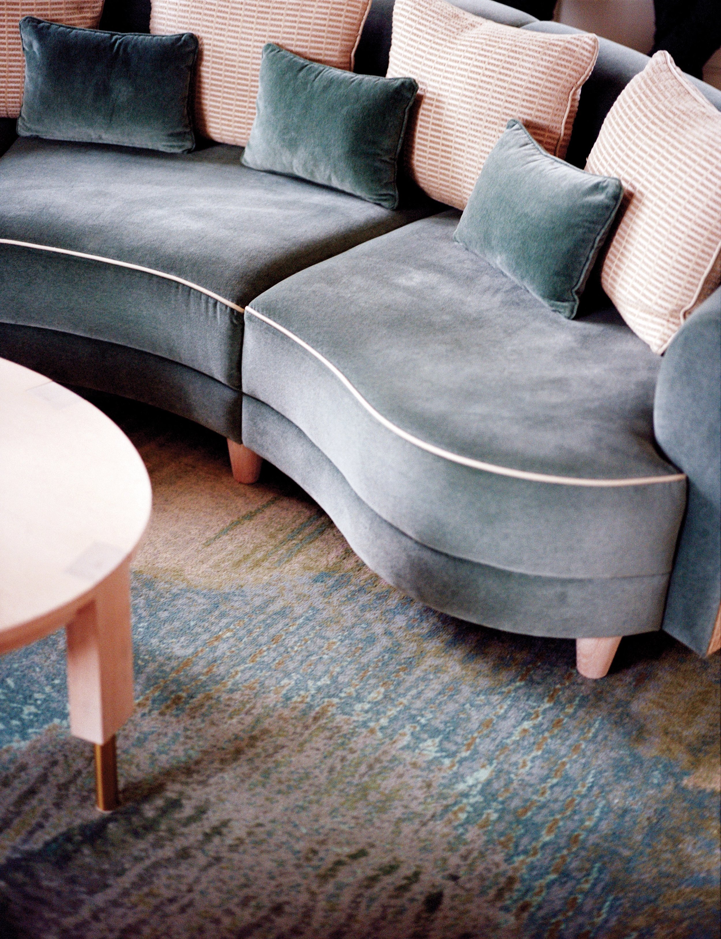 Close-up view of a sofa with throw pillows, some solid dark green and some with a pink geometric pattern, placed against a multicolored rug and a corner of a light-colored wooden side table.
