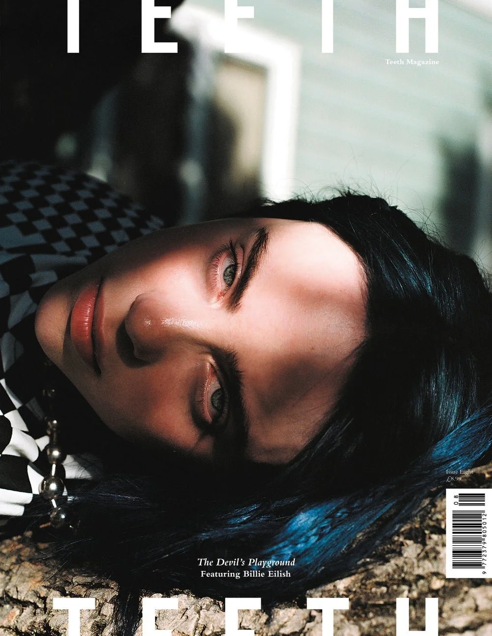 Close-up of a woman with black hair, lying down outdoors, with sunlight on her face, wearing a black and white checkered shirt and a bead necklace, on the cover of a magazine.