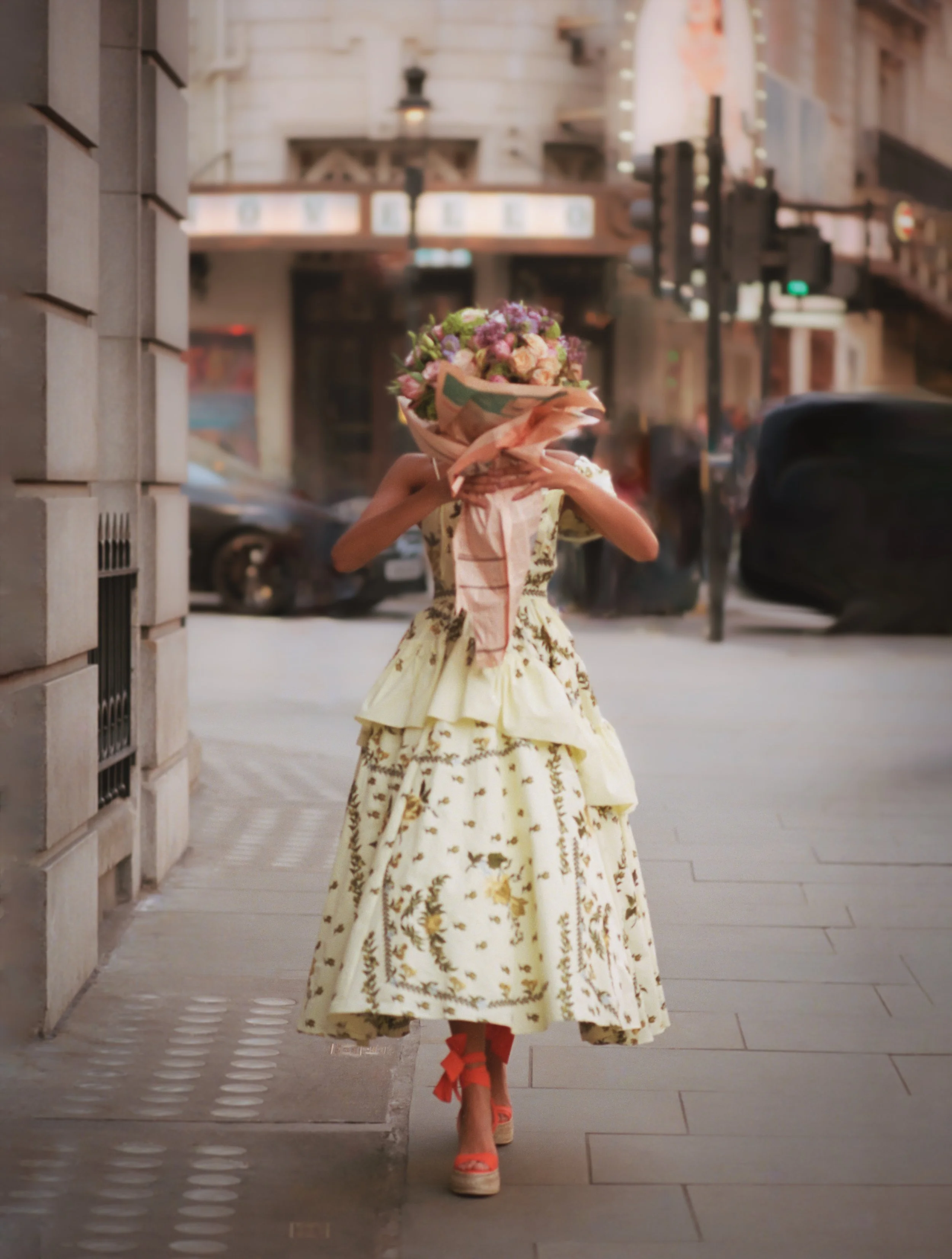 A woman in a vintage yellow floral dress walking on an urban sidewalk, holding a large bouquet of flowers in front of her face, with only her red high heels visible.