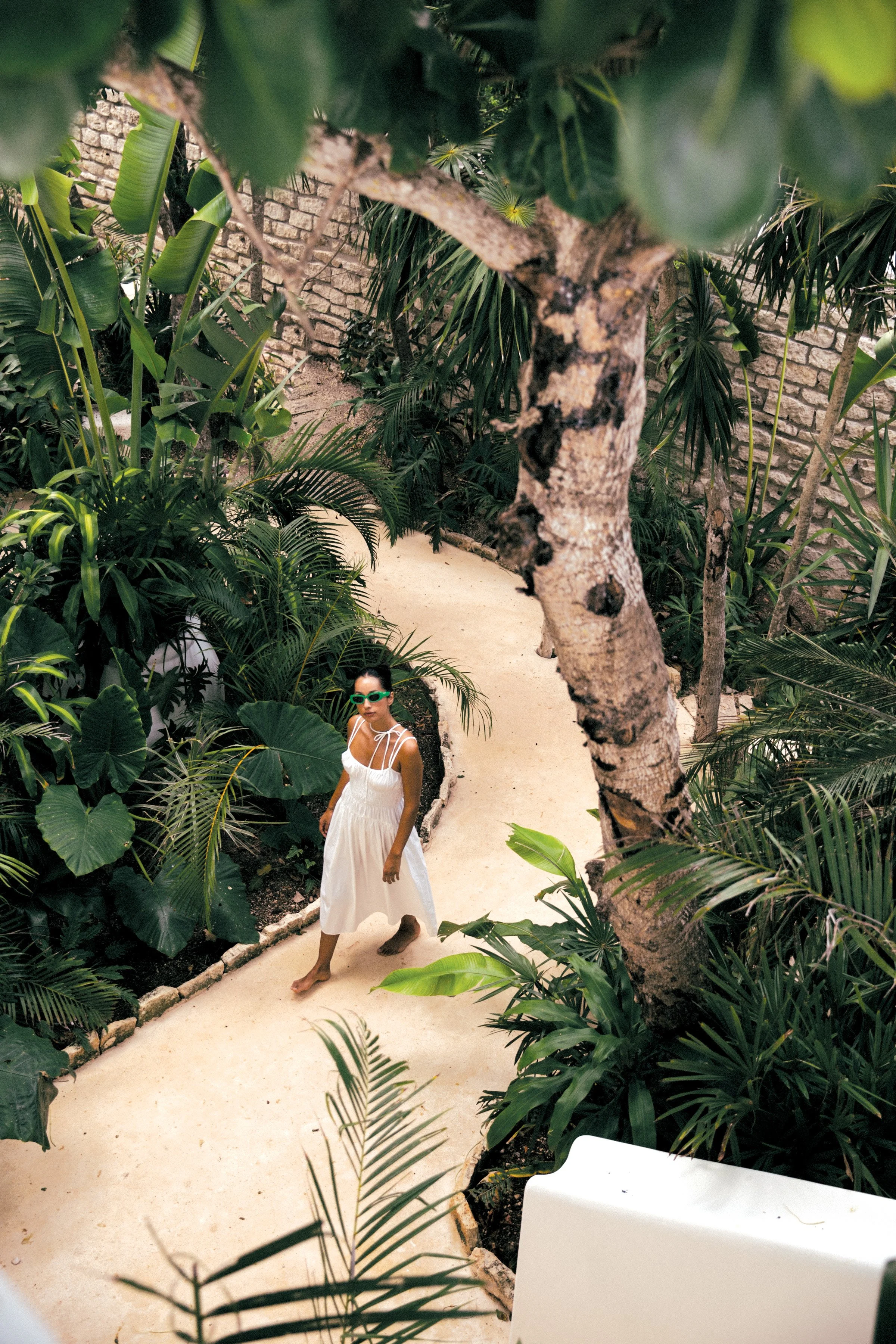 A woman in a white dress and green sunglasses walking barefoot on a light-colored pathway surrounded by lush green tropical plants and trees, with a brick wall in the background.