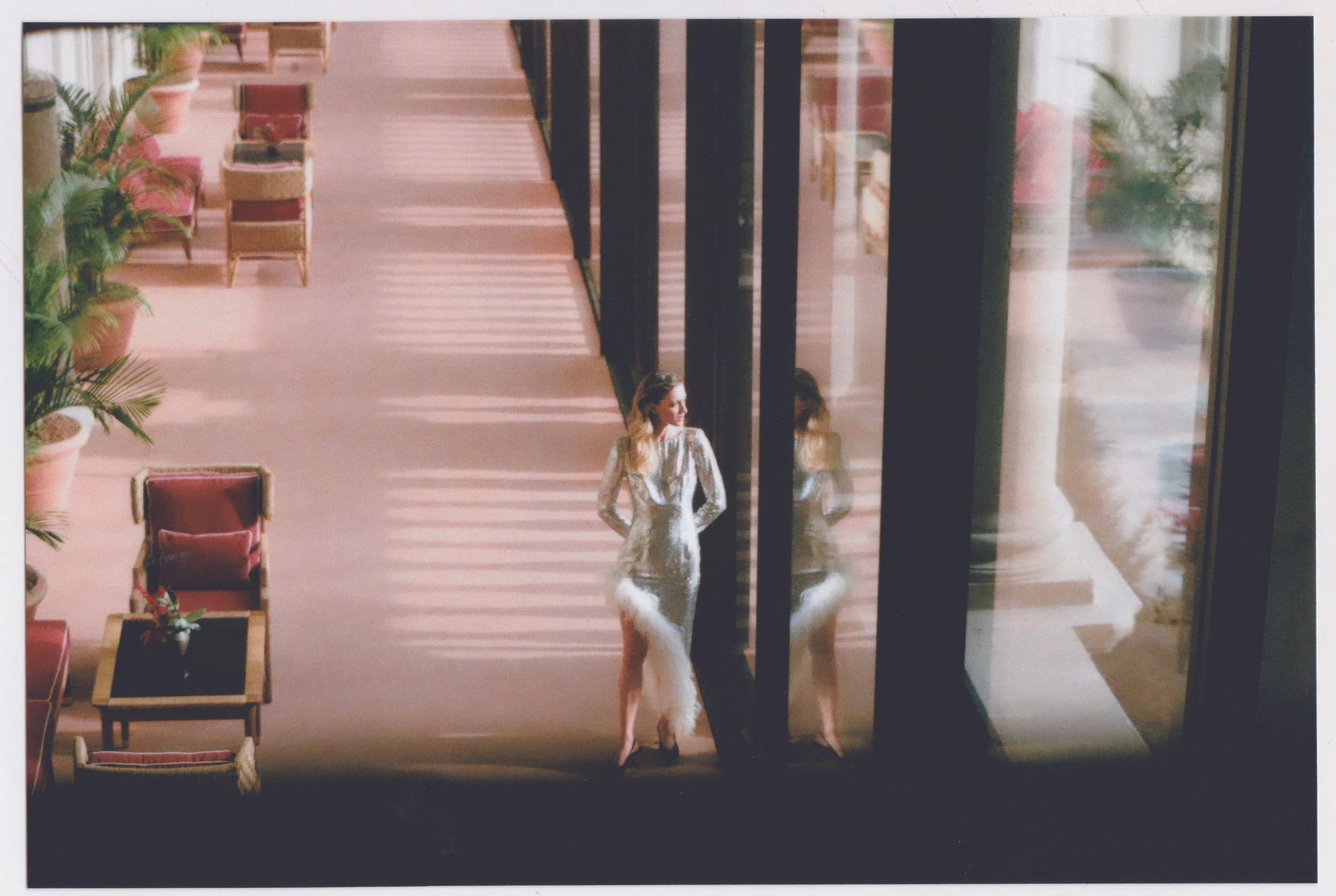 A woman in an elegant dress stands on a wooden floor near glass doors, with her reflection visible. Potted plants and chairs are arranged along the corridor.