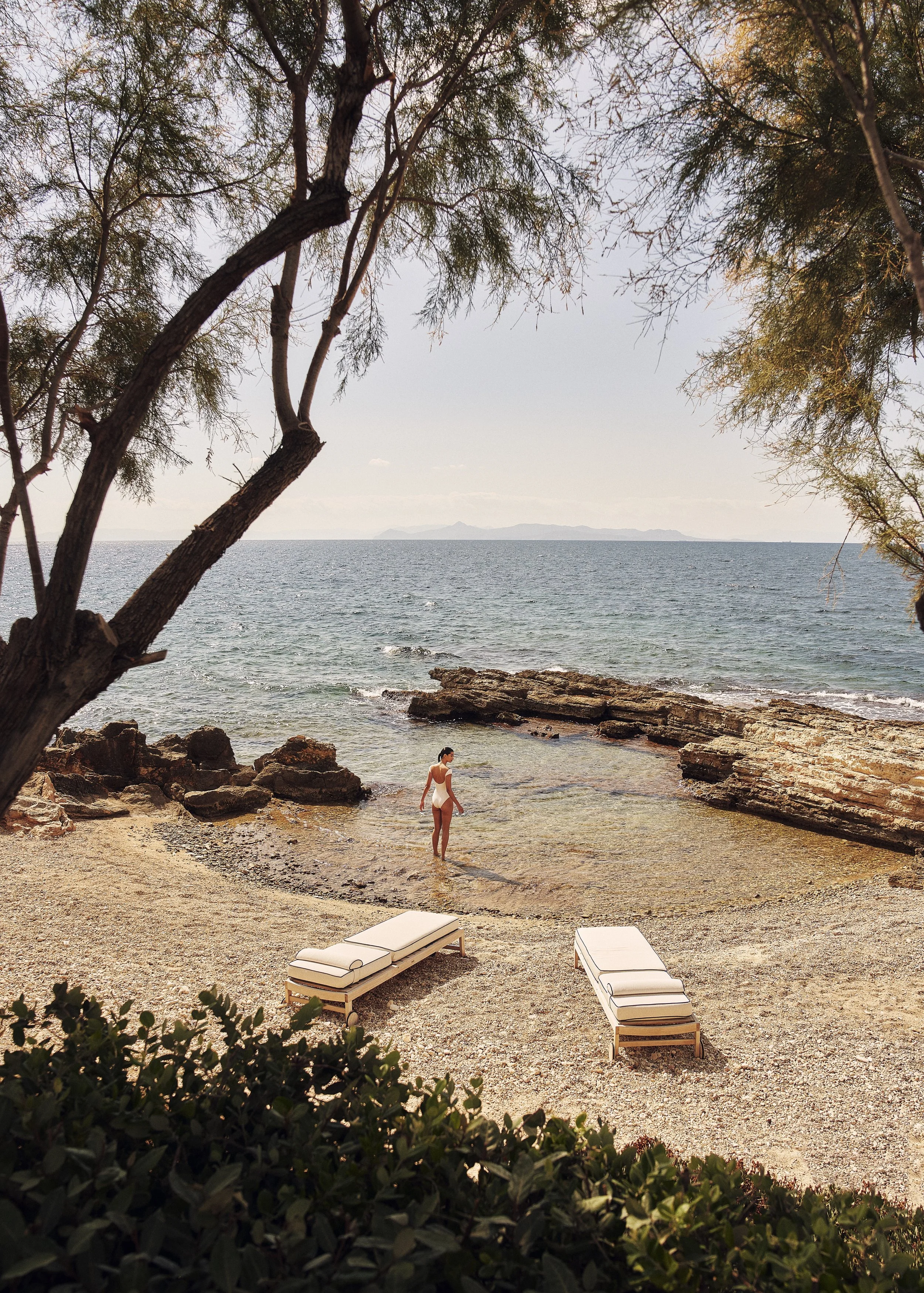 A woman in a bikini walking into the ocean on a rocky beach, framed by trees and lounge chairs in the foreground, with distant islands on the horizon under a partly cloudy sky.