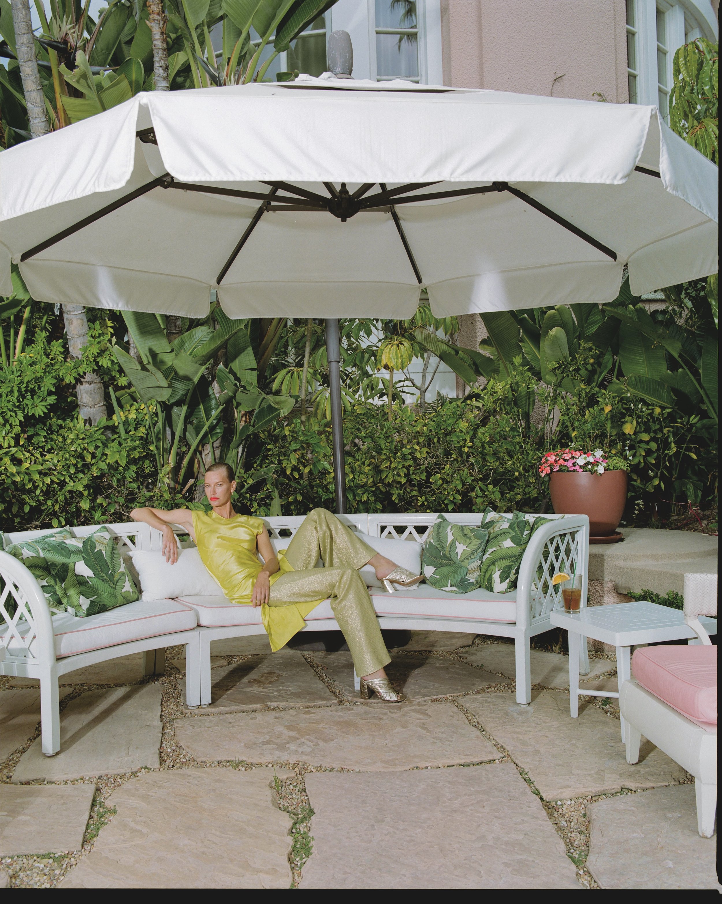 Woman in yellow shiny outfit and metallic high heels sitting on a white outdoor sofa under a large white patio umbrella, surrounded by greenery and potted plants.