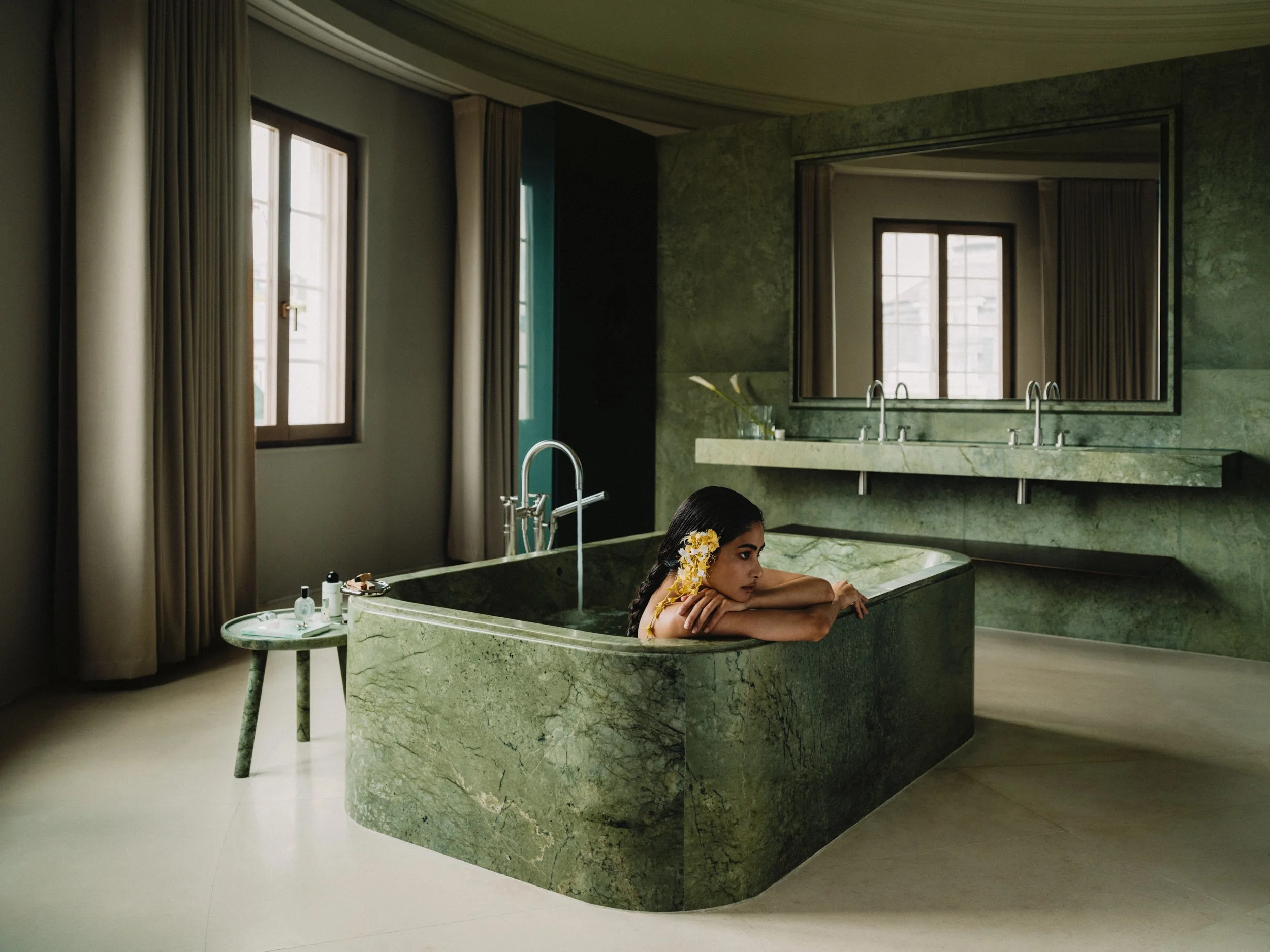 A woman relaxing in a green marble bathtub in a modern bathroom with large windows and beige curtains.