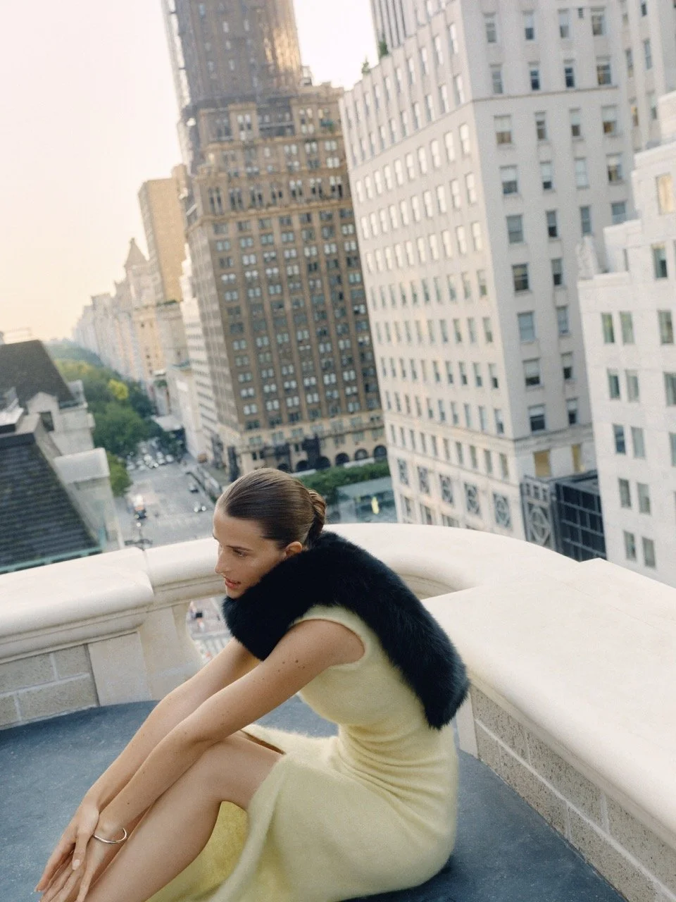 A woman wearing a light yellow dress and black fur stole sits on a balcony overlooking a city street, with tall buildings in the background.