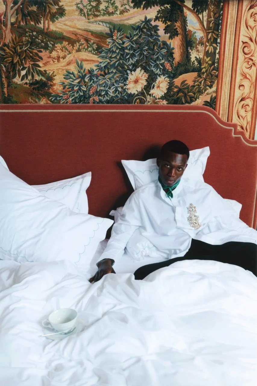 A young man with dark skin and short hair lying on a bed with white bedding, wearing a white shirt with a decorative emblem on the chest, and a green neckerchief. He is sitting up against a red headboard, with a teacup and saucer placed on the bed in