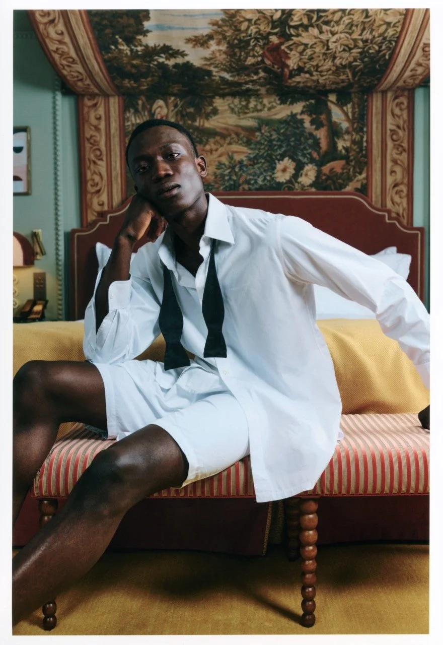 A man sitting on a bed in a vintage-style room, wearing a white shirt and shorts, with a loosened black tie, resting his head on his hand, in front of a floral tapestry headboard.