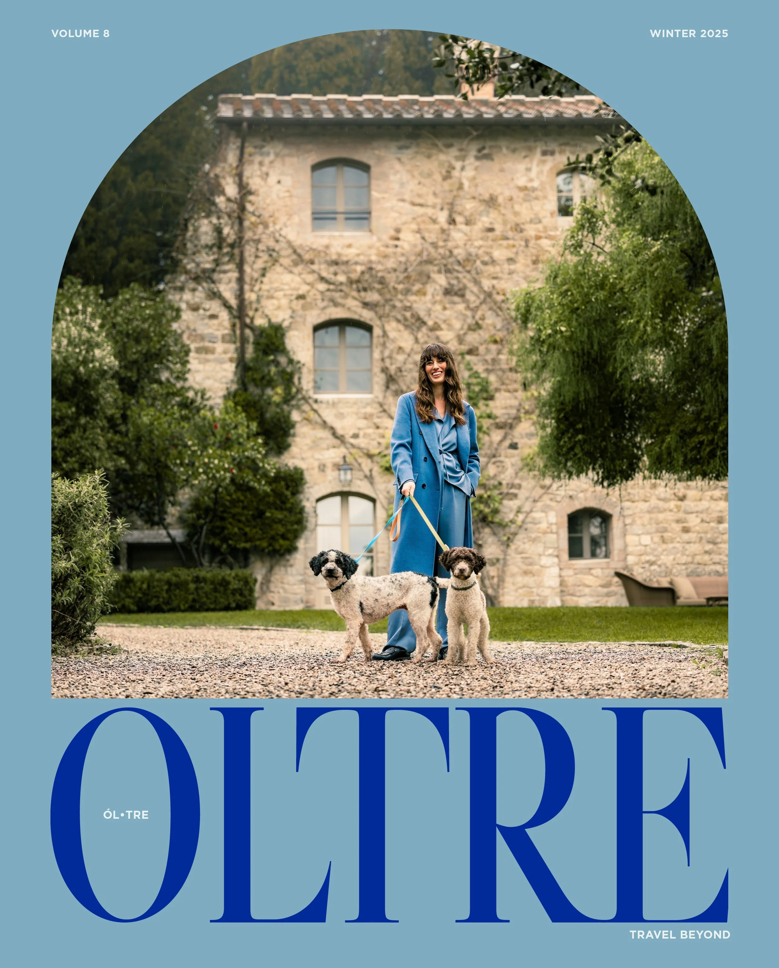 A woman in a blue coat smiling while walking two dogs on a leash in front of an old stone building with multiple arched windows, surrounded by greenery during daytime.