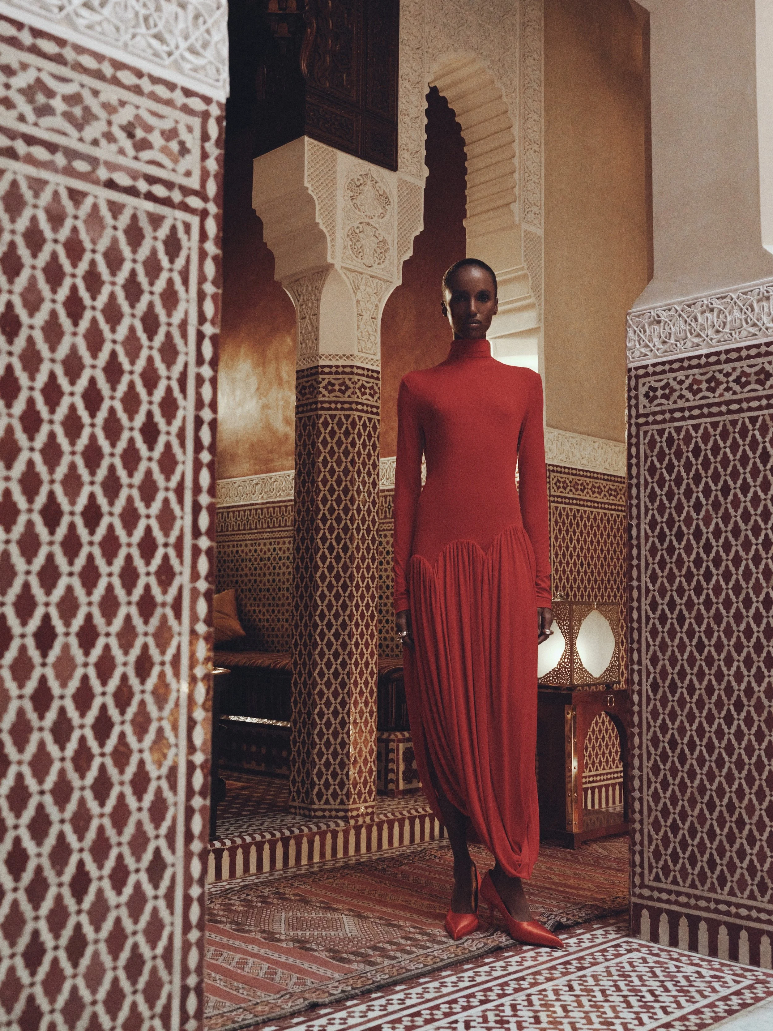 A woman dressed in a long, elegant red dress and red high heels standing in a Moroccan-style room with intricate white and red tile patterns, ornate arches, and decorative screens.