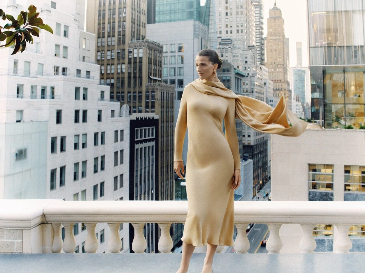A woman in a beige dress with an attached scarf standing on a balcony overlooking a cityscape of tall buildings.