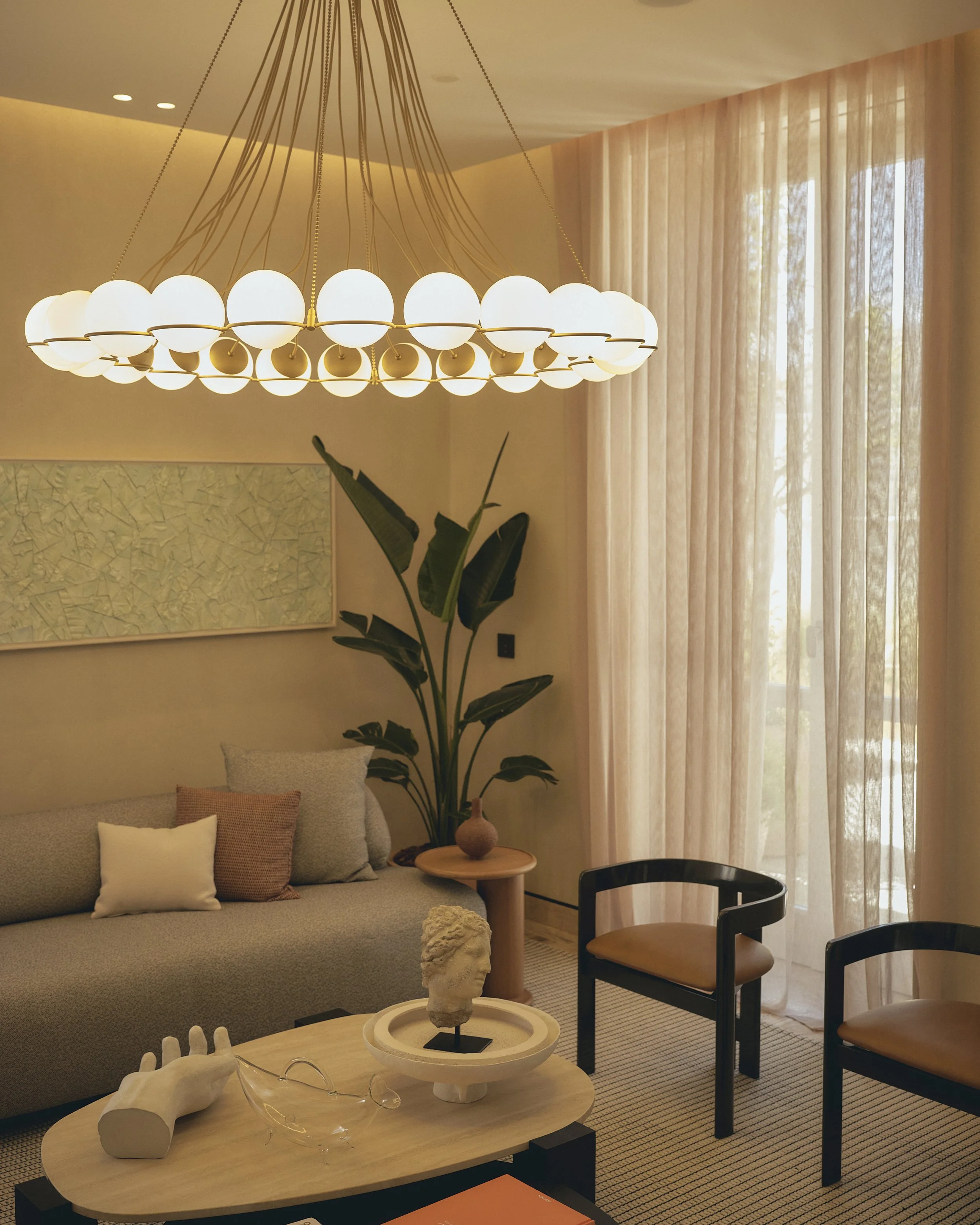 Living room with beige sofa decorated with pillows, a large plant near the window with sheer curtains, a wooden coffee table with a white sculpture, glasses, and a bright chandelier hanging from the ceiling.