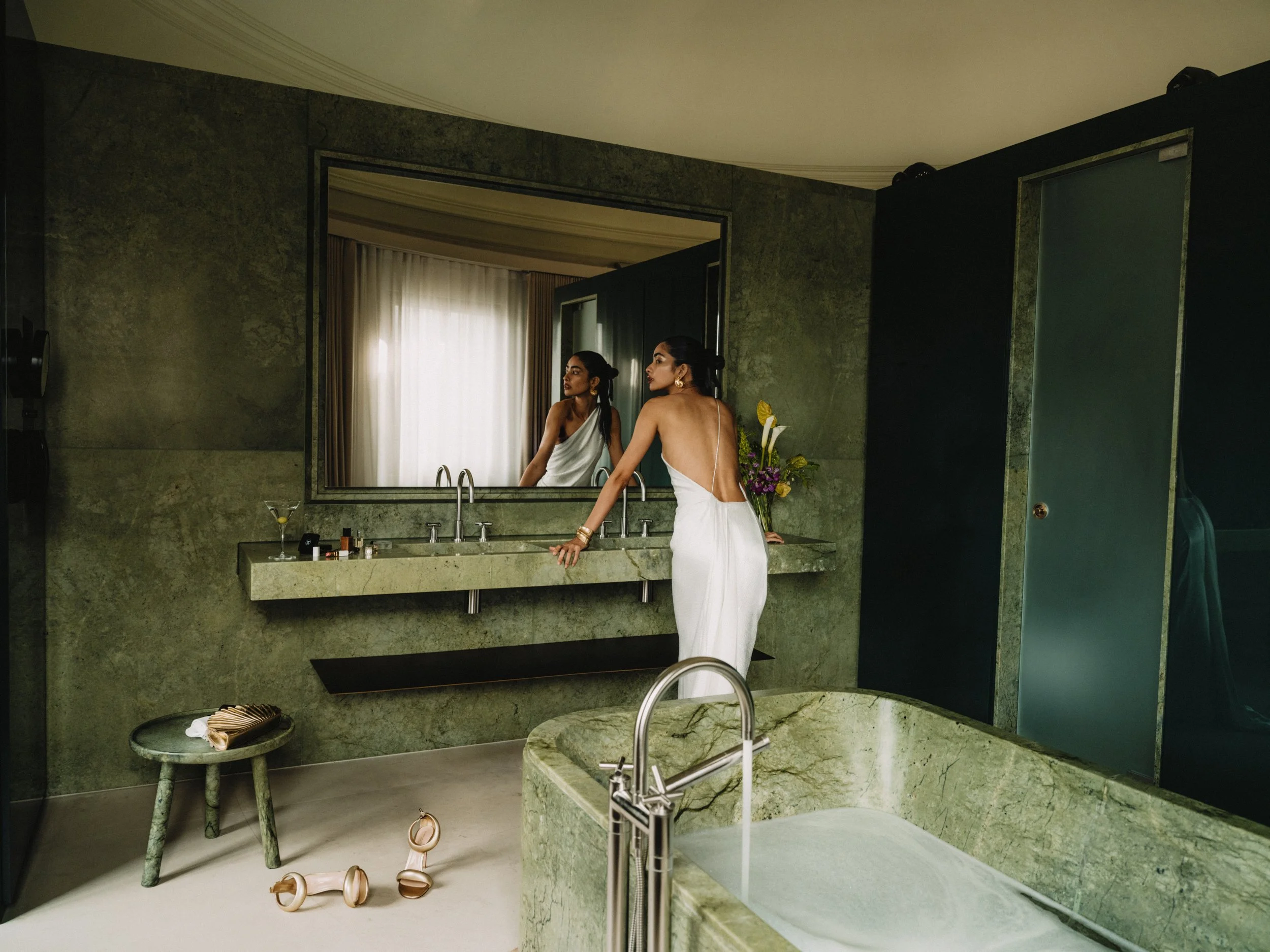 A woman in a white towel with gold jewelry stands in a stylish bathroom looking at herself in a mirror, with pink high-heeled shoes on the floor nearby and a bouquet of flowers on the counter.