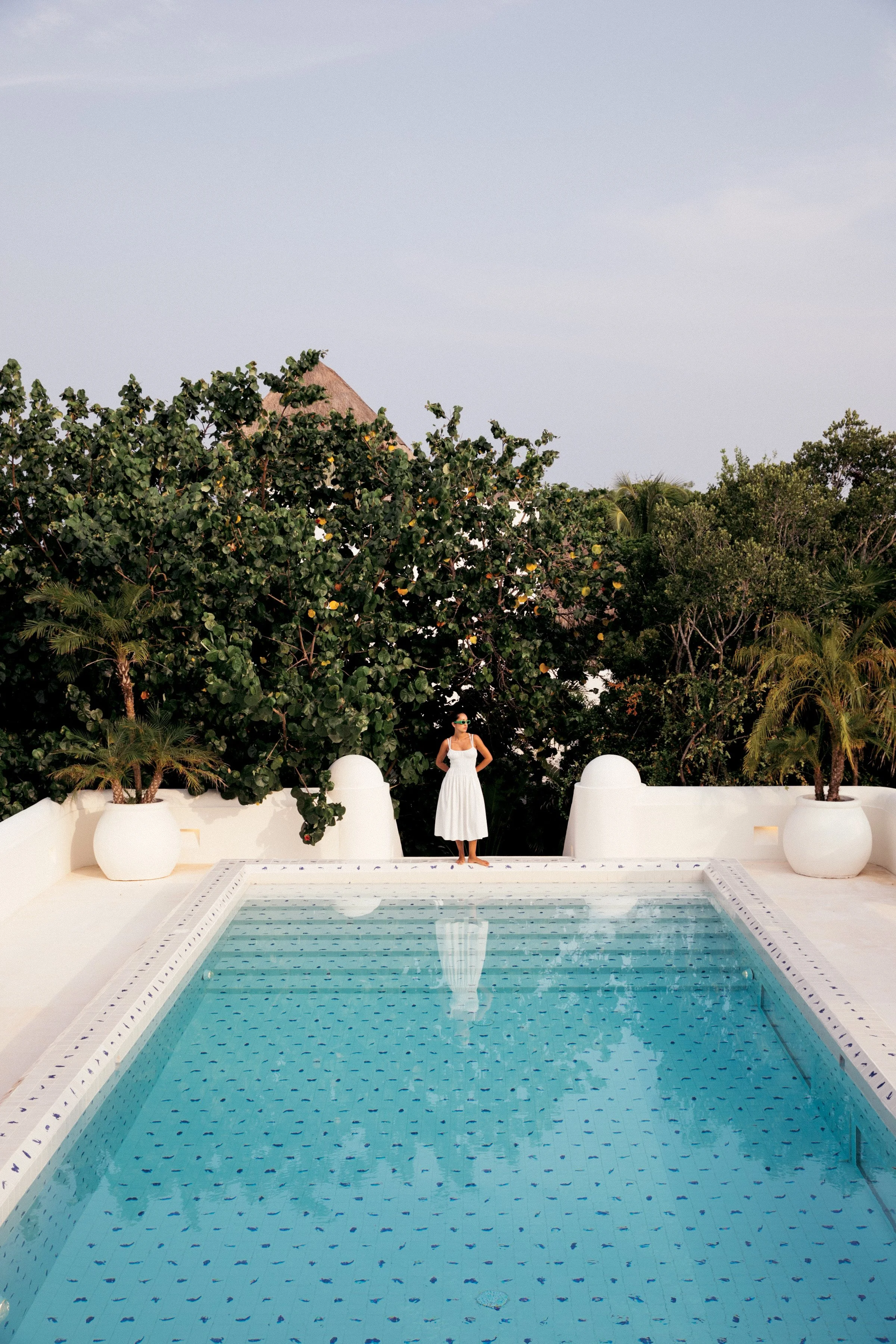 A woman in a white dress standing at the edge of a swimming pool with clear water, surrounded by white walls and large potted plants, with lush trees and a thatched roof structure in the background under a cloudy sky.