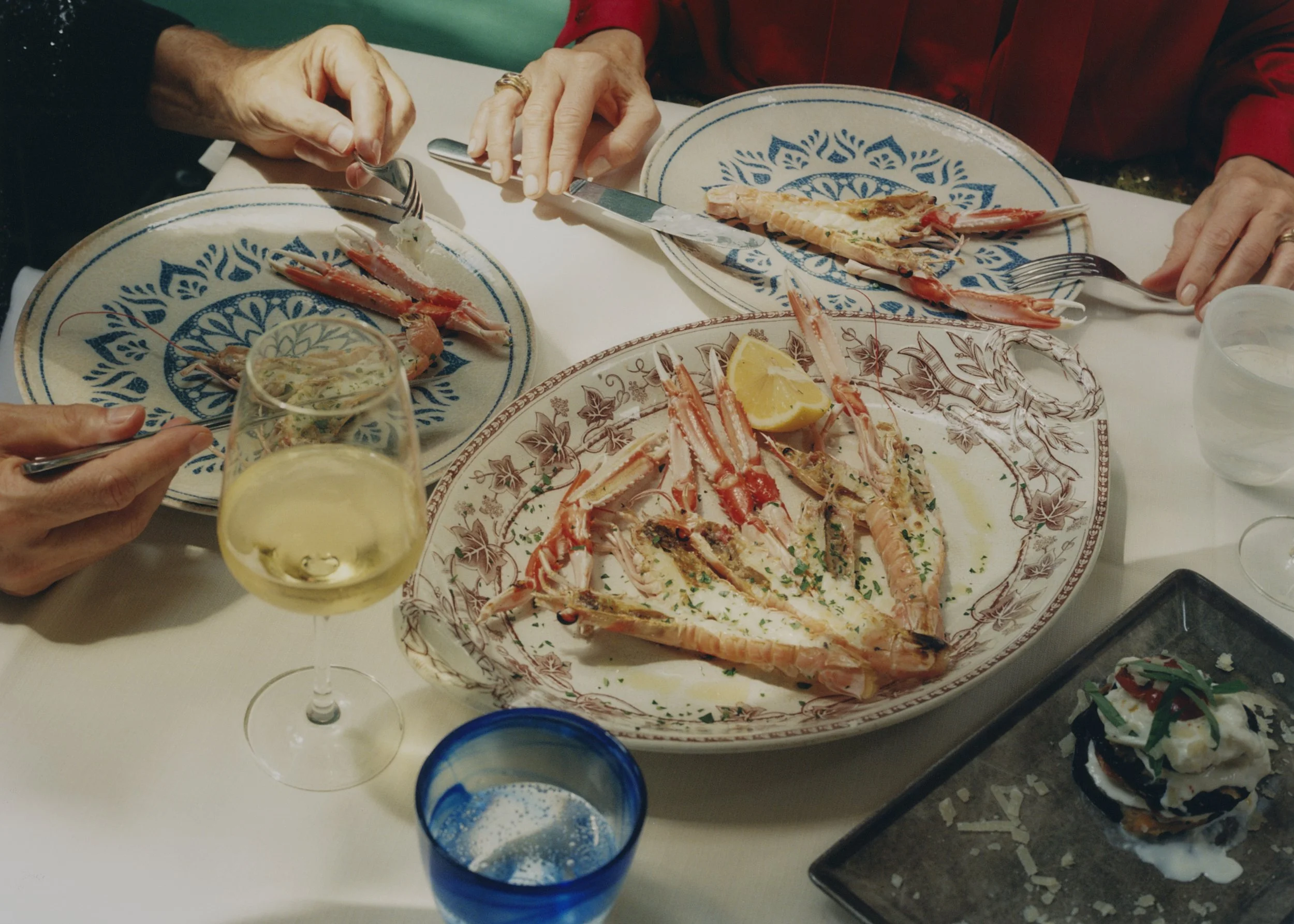 A dinner scene with two plates of lobster tails, a lemon wedge, and a glass of white wine on a white table. Two people are cutting and eating lobster, with one person's hands visible, one wearing a black long sleeve and the other in red.