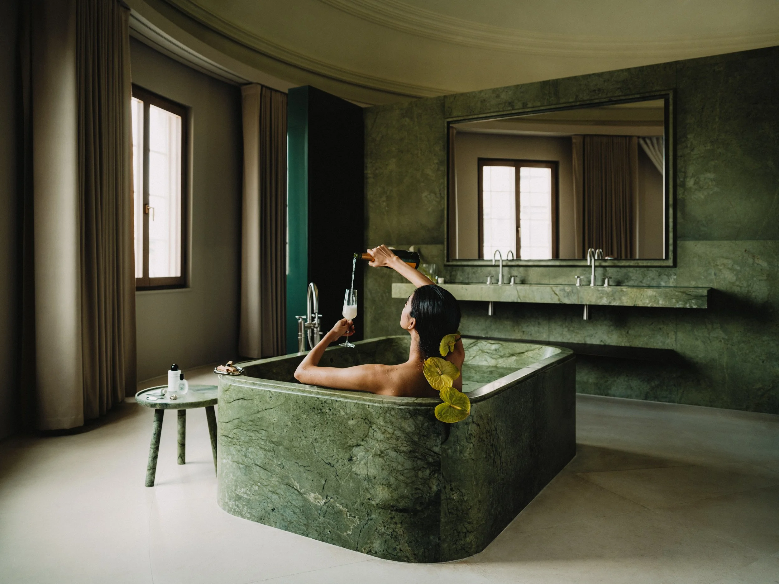 A woman is relaxing in a green marble bathtub, holding a glass of champagne, with a large rectangular mirror and windows with beige curtains in the background.