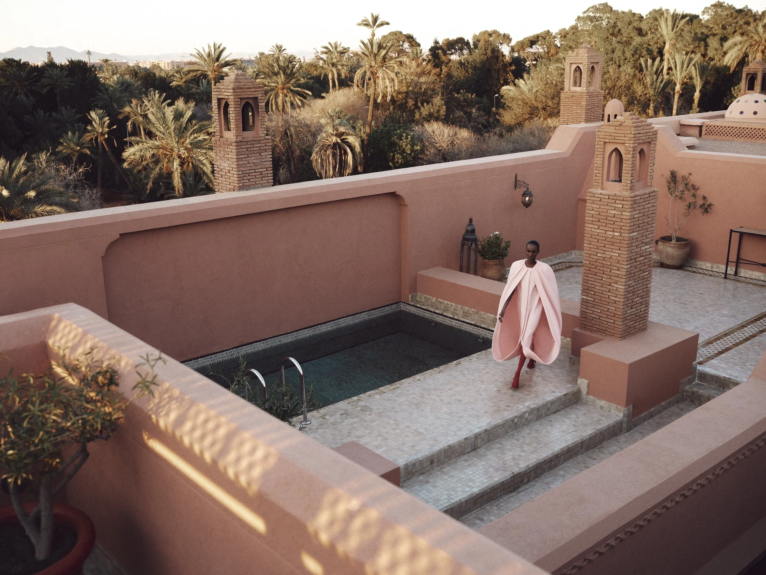 A woman walking on a pink courtyard with a pool, surrounded by desert plants and palm trees, with a pink wall and chimneys in the background.