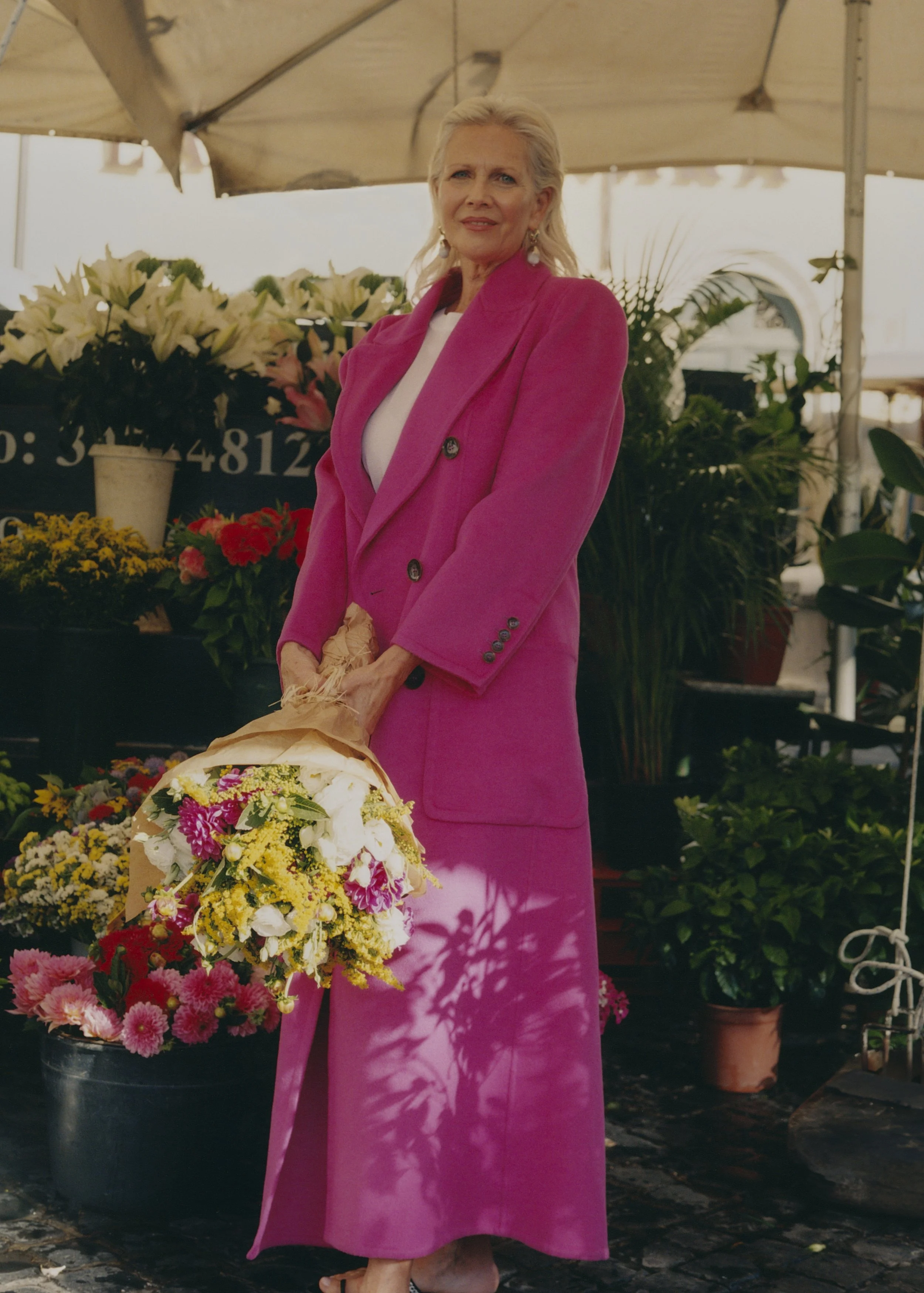 A woman with blonde hair dressed in a bright pink suit standing outdoors at a flower market, surrounded by various potted plants and flower arrangements.