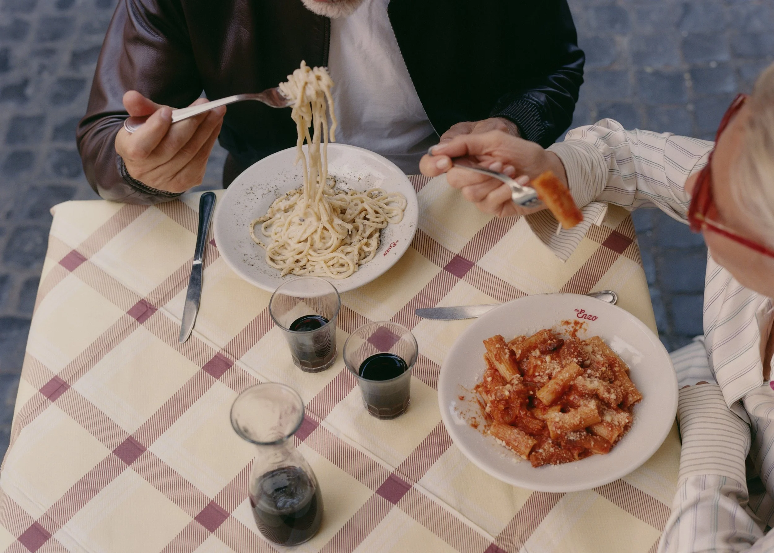 Two people sharing a meal at a restaurant table with spaghetti, kimchi, and glasses of red wine.