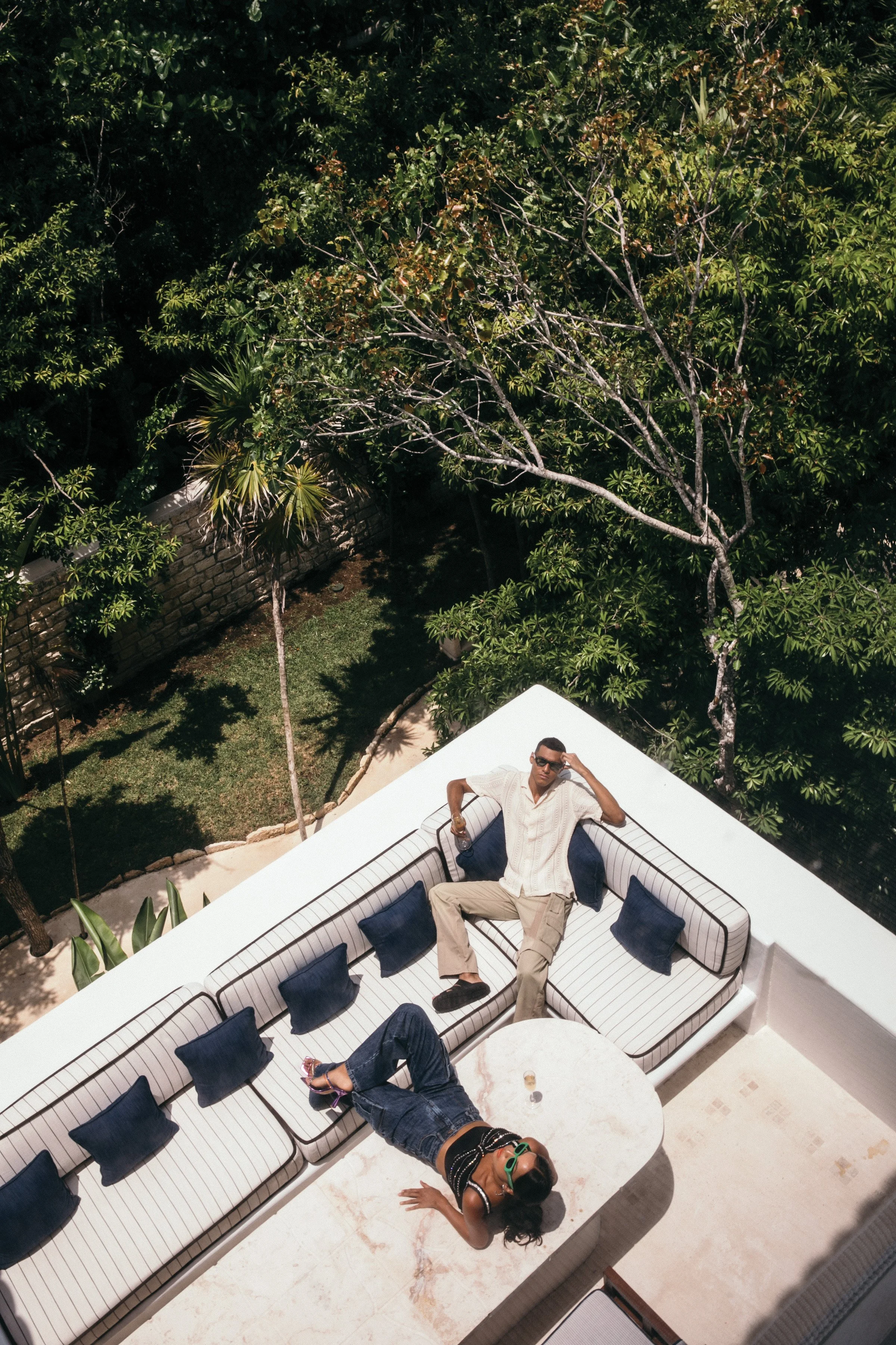 Two people relaxing on a outdoor white striped sofa with blue pillows, one man sitting and one woman lying on her back on a table, on a balcony with greenery and trees in the background.
