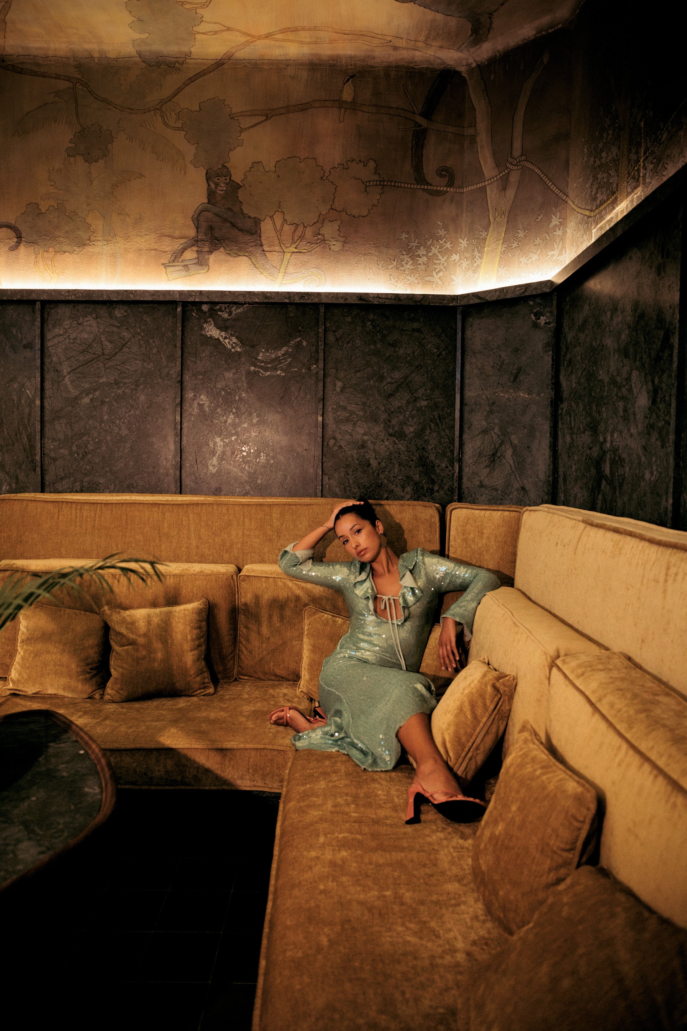 A woman in a shiny, sequined dress with a slit, sitting on a yellow sofa with matching cushions in a dimly lit room. The room has dark marble walls and a mural of a monkey in the trees painted on the ceiling in the background.