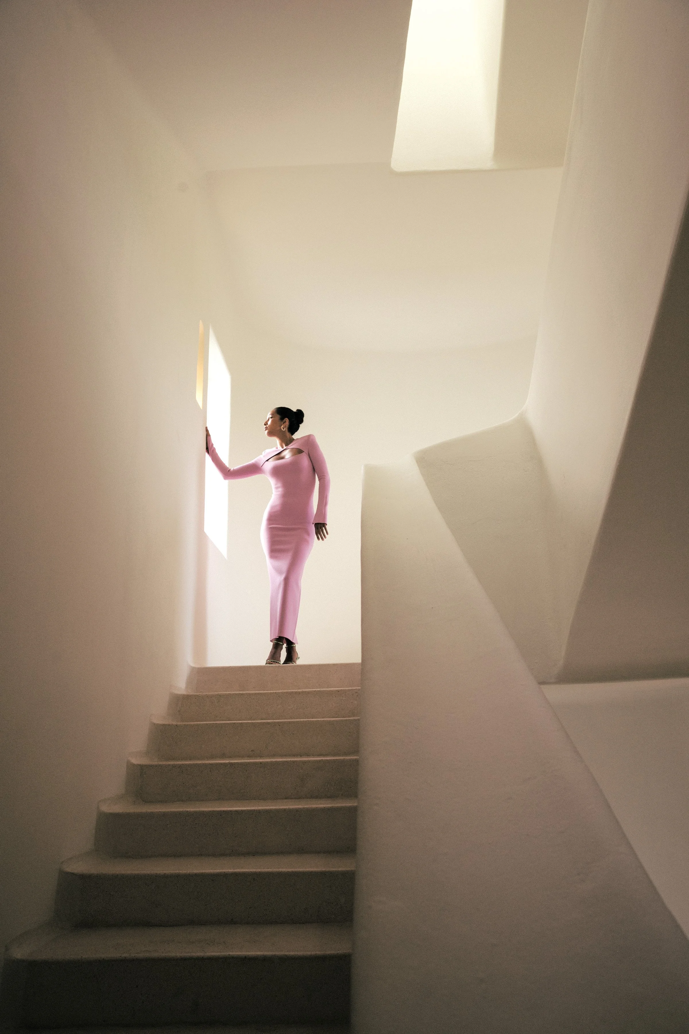 A woman in a long pink dress stands on stairs near a window, with her hand resting on the window frame, in a modern minimalistic interior with curved white walls.
