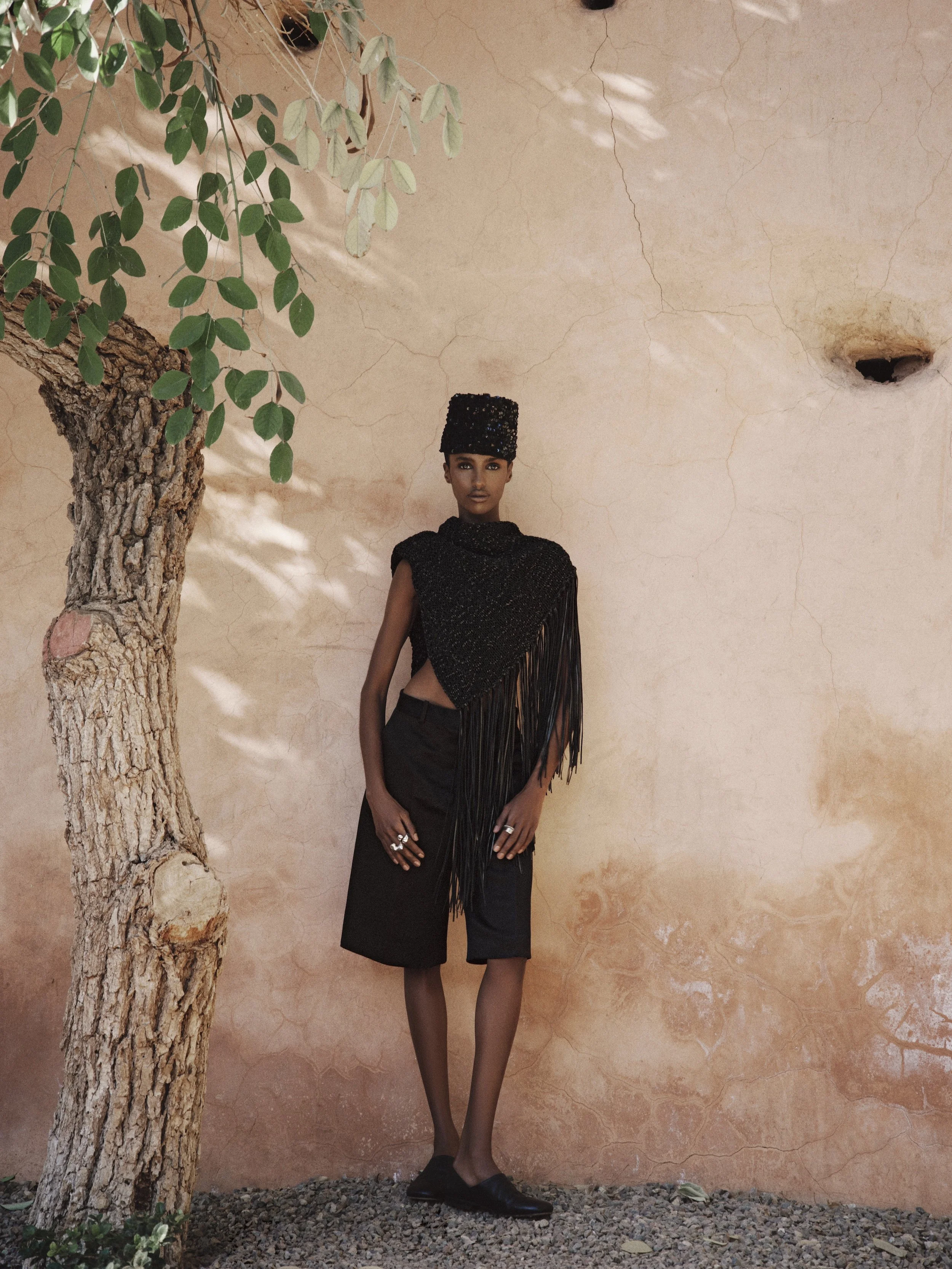 A person standing next to a tree and a beige stucco wall, wearing a black textured top with fringe, black wide-leg pants, black shoes, and a black beaded hat.
