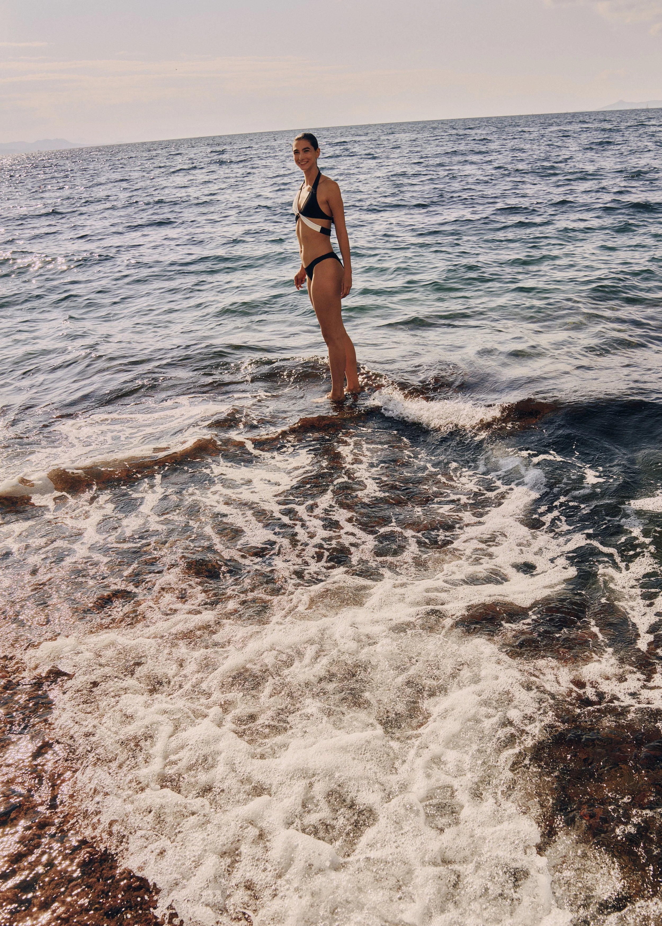 A woman in a black bikini standing in the shallow water at the beach with the ocean in the background and a cloudy sky.
