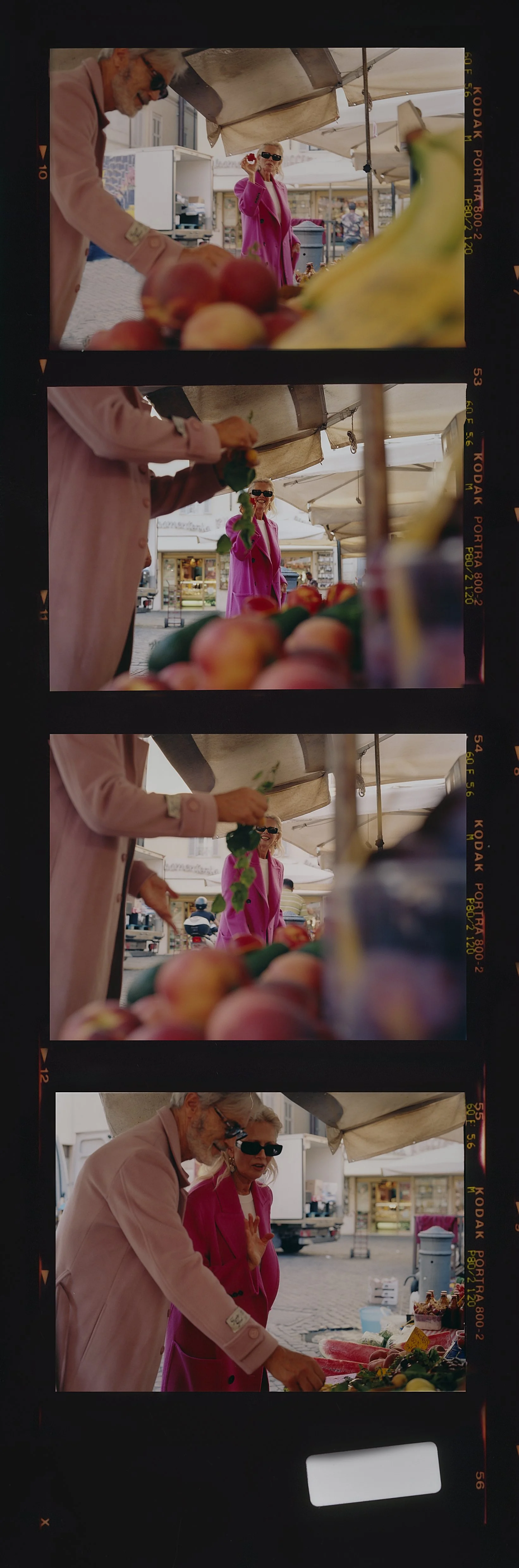 Two women shopping at an outdoor market, one in a pink coat and black sunglasses, the other in a light pink jacket, browsing produce including peaches, bananas, and cucumbers.
