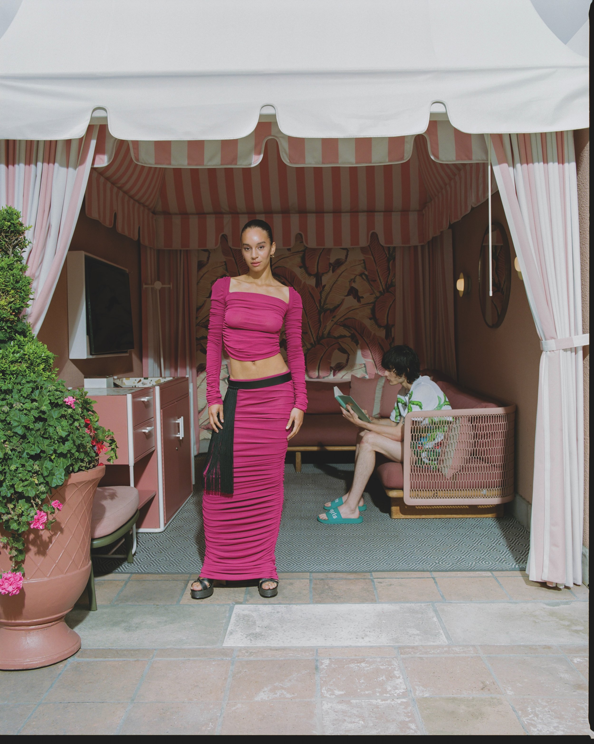 A woman in a pink long-sleeve dress with black pleated skirt standing in front of a pink-striped cabana with a tropical leaf mural inside, a man sitting on a pink bench reading a book, and a large potted plant with pink flowers on the left.